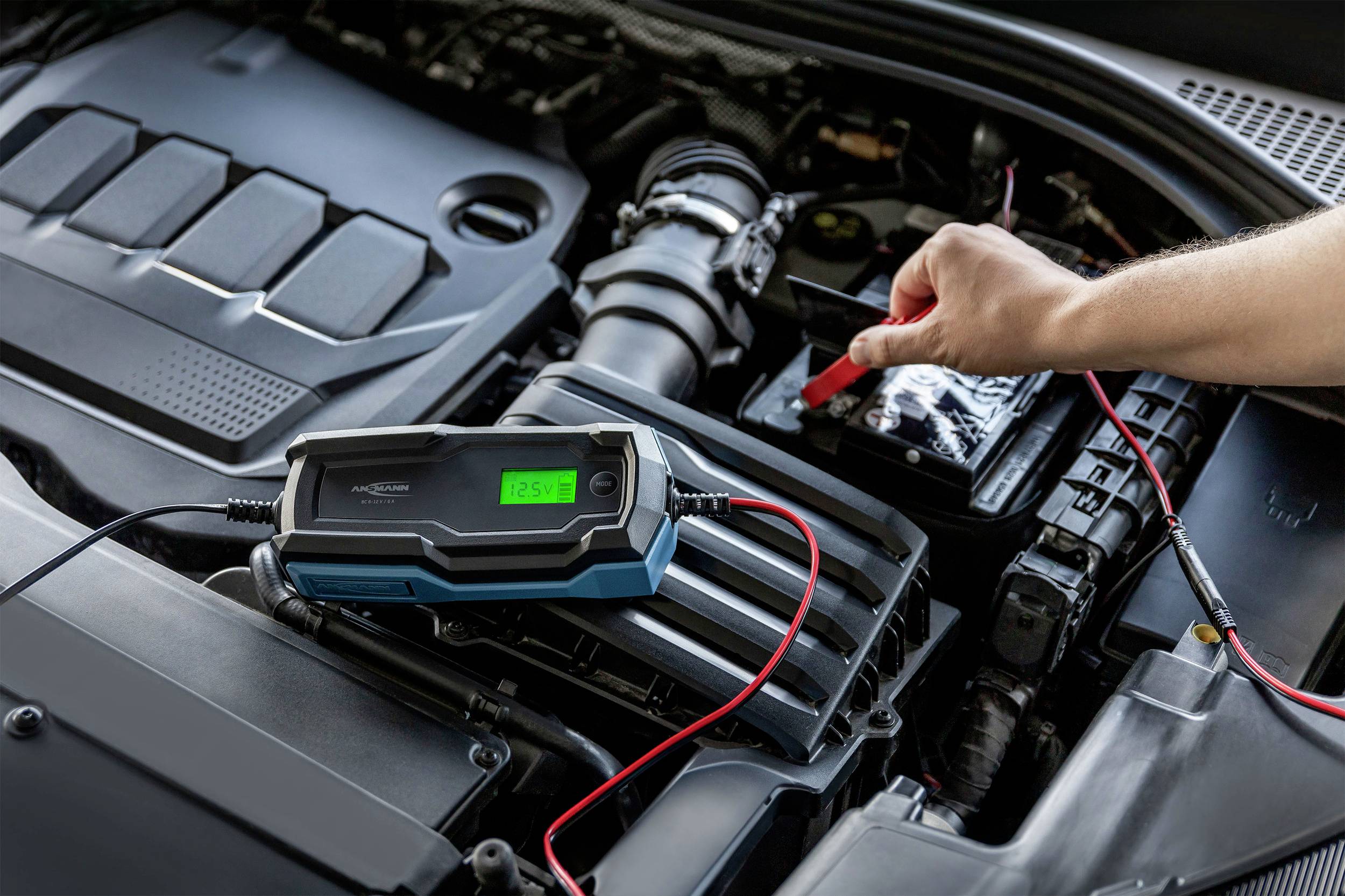 Eine Hand schließt ein Kabel eines Batterieladegeräts an eine Autobatterie an. Das Display des Ladegeräts zeigt 12,5V an.