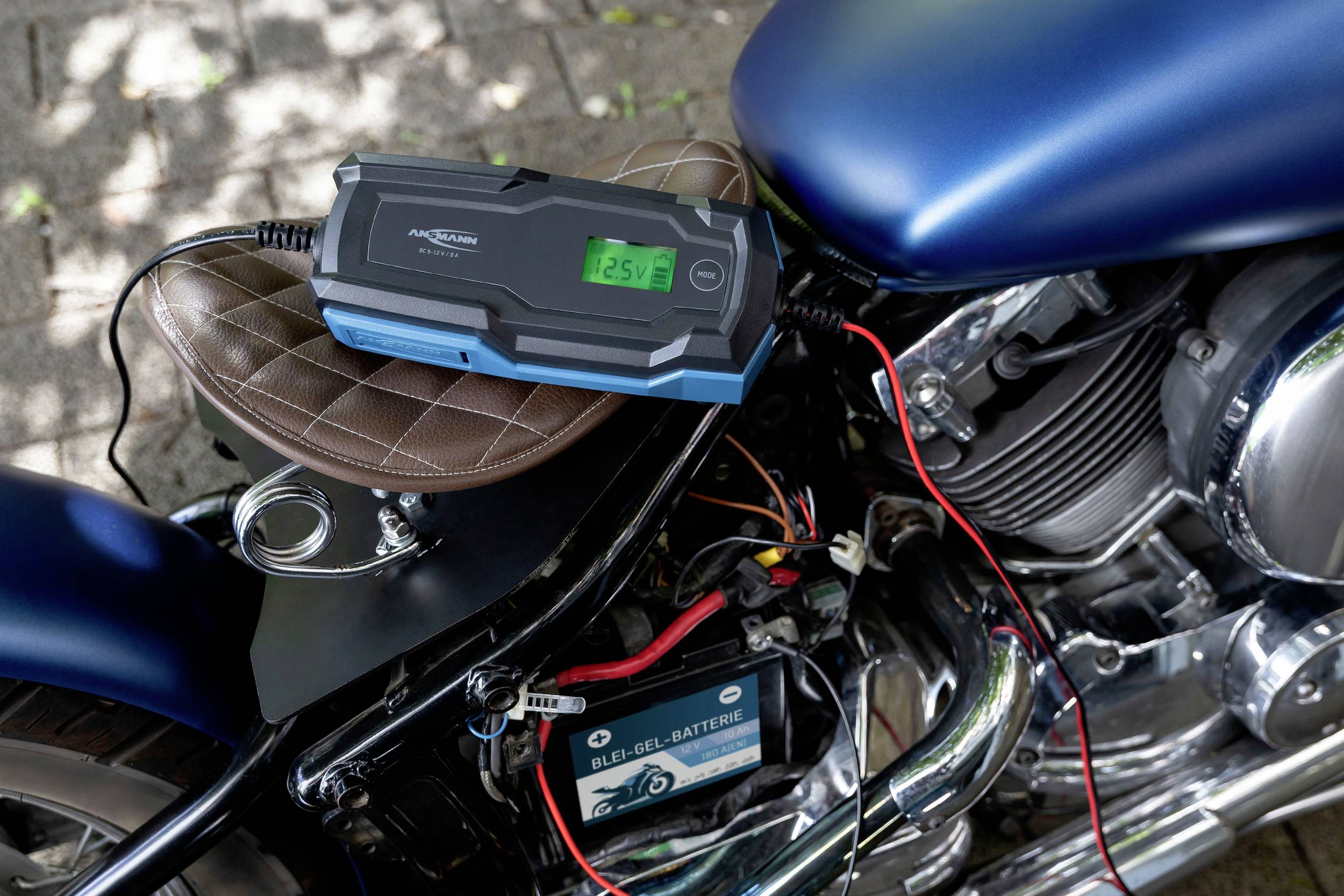 Motorrad steht auf einem Gehweg, verbunden mit einem Batterieladegerät. Das Display zeigt '12.5V' an. Schatten von Bäumen im Hintergrund.