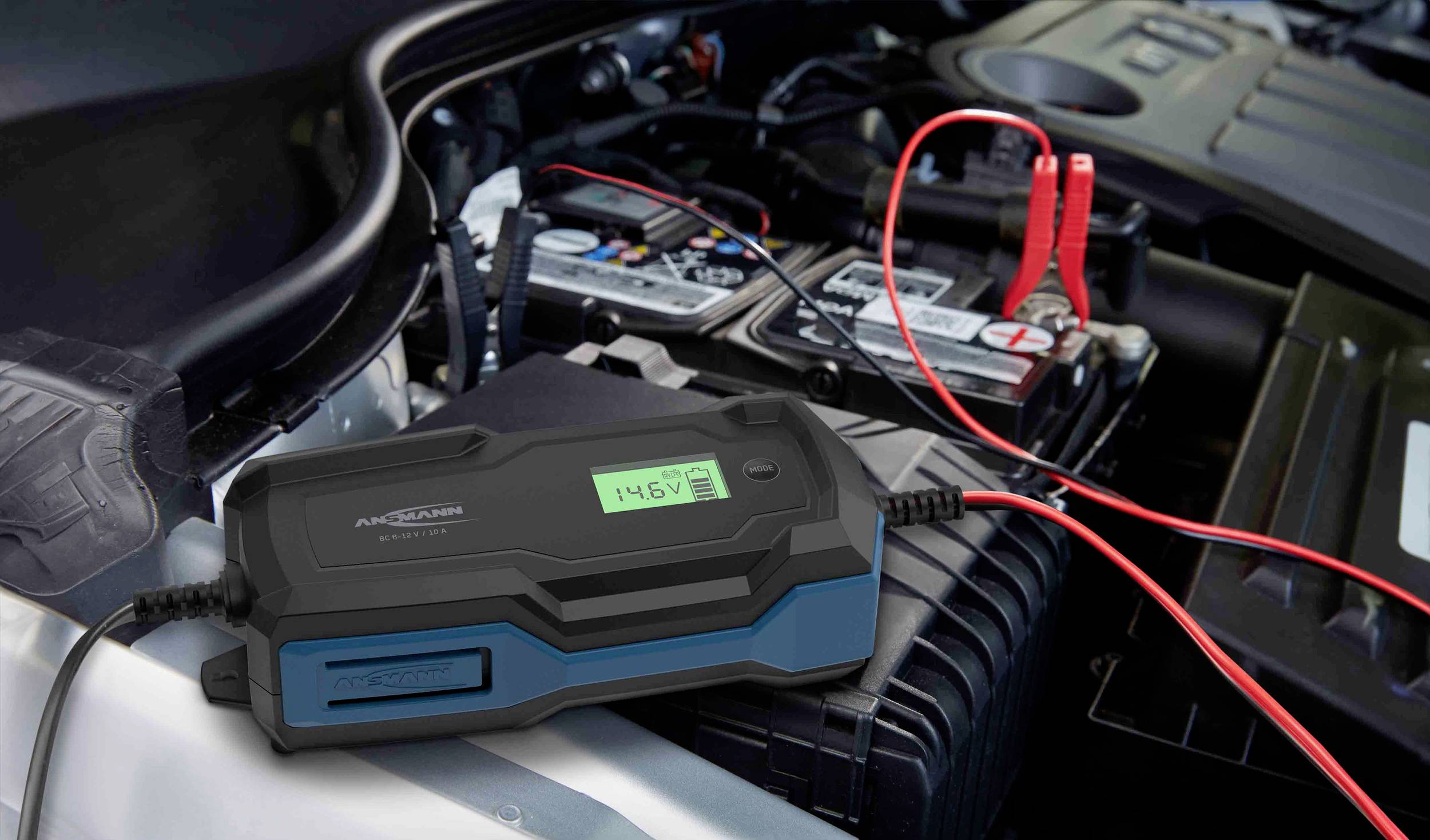 Ein Auto mit geöffneter Motorhaube zeigt ein Batterieladegerät, das an die Autobatterie angeschlossen ist. Ein Display zeigt '14,6V'.