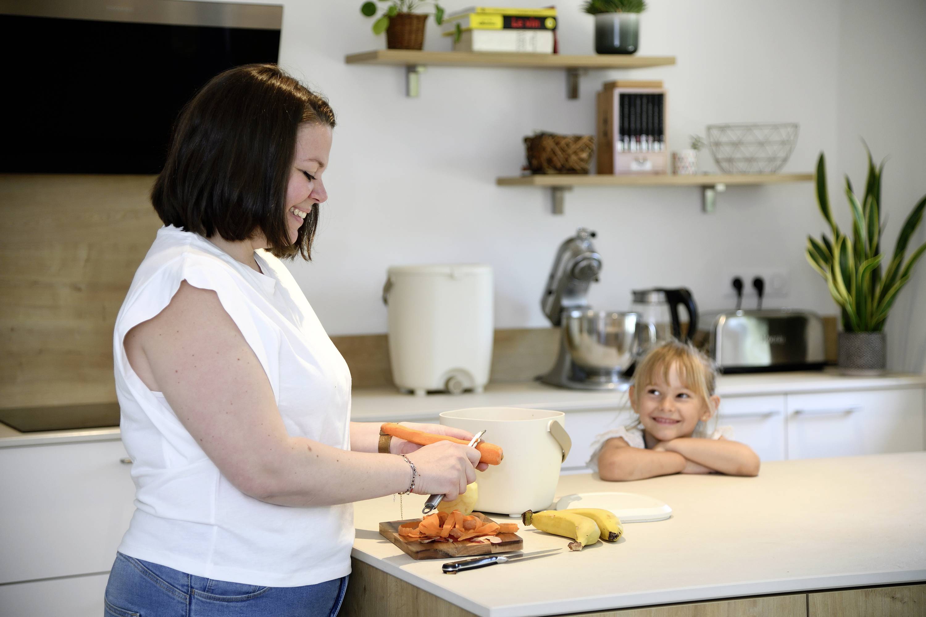 Eine Frau schält Karotten in einer modernen Küche, während ein Kind lächelnd am Tisch sitzt und zuschaut.