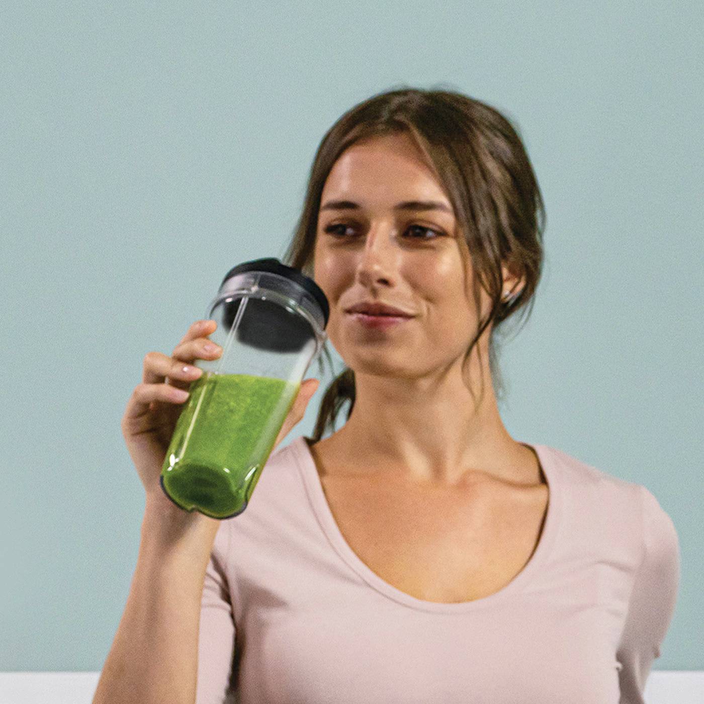 Eine Frau hält einen Becher mit grünem Smoothie und lächelt. Sie trägt ein rosa Oberteil und hat langes Haar. Hintergrund ist neutral.