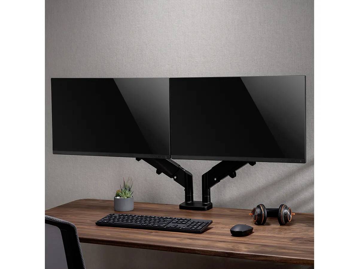 Doppelter Monitorarm hält zwei Bildschirme über einem Holztisch mit Tastatur, Maus, Kopfhörer und Pflanze.