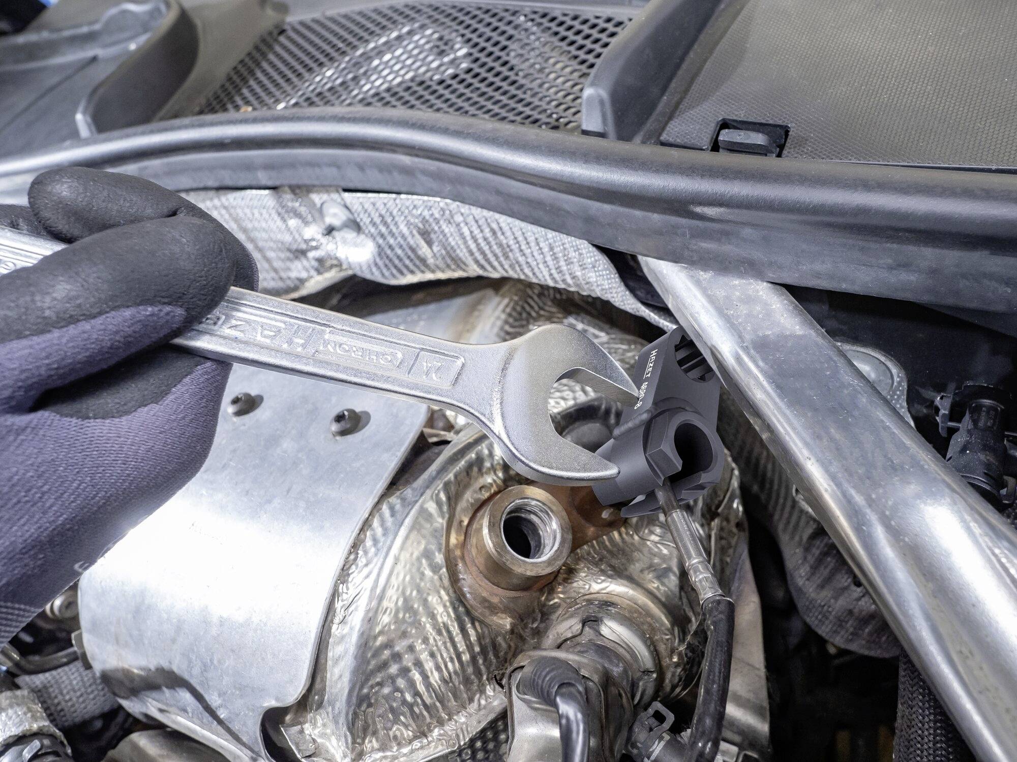 Ein Mechaniker hält einen Schraubenschlüssel im Motorraum eines Autos. Der Schraubenschlüssel greift eine Mutter in einem komplexen Teil.