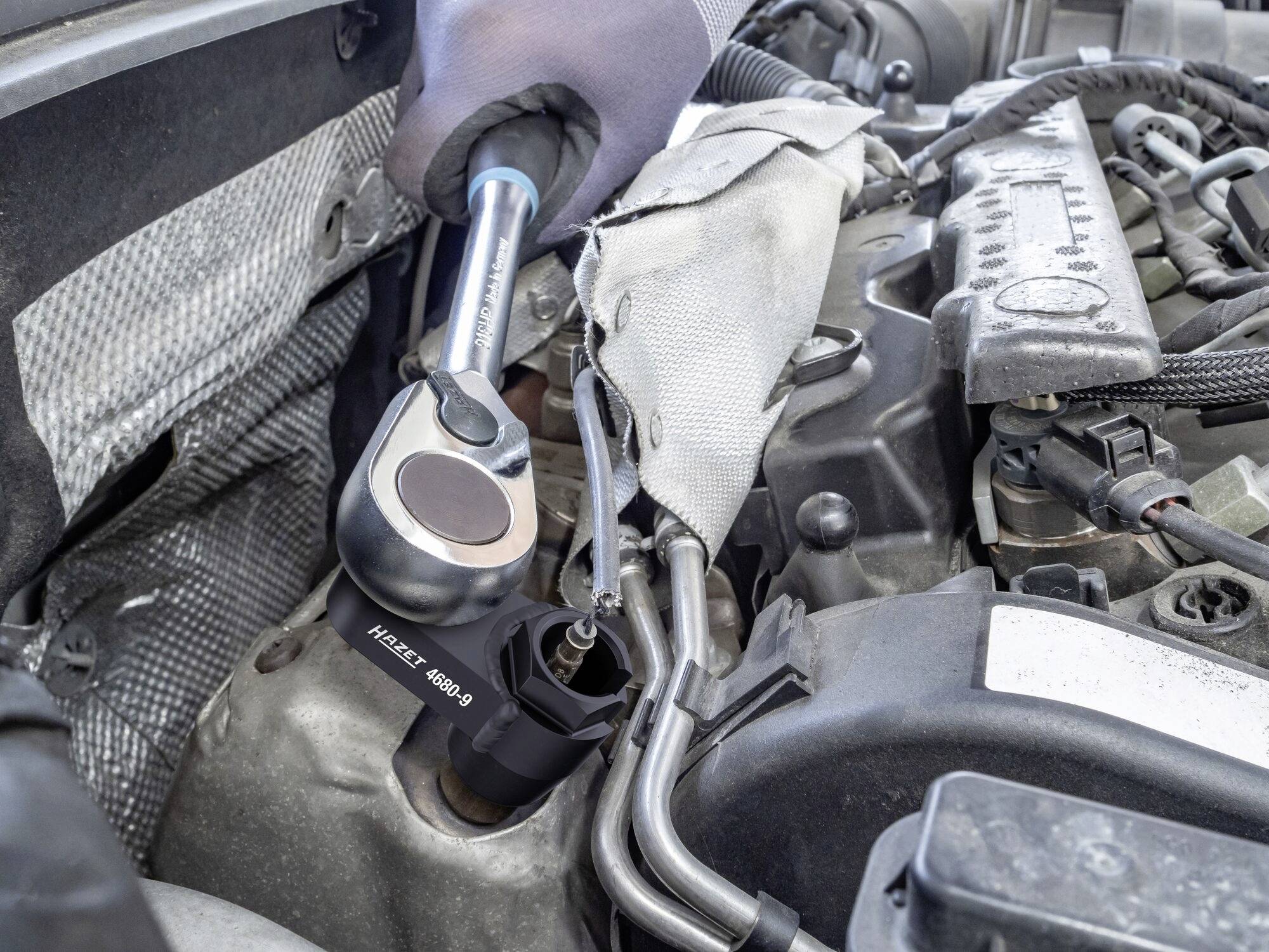 Ein Mechaniker zieht mit einem Drehmomentschlüssel eine Schraube im Motorraum eines Autos an. Verschiedene Motorbauteile sind sichtbar.