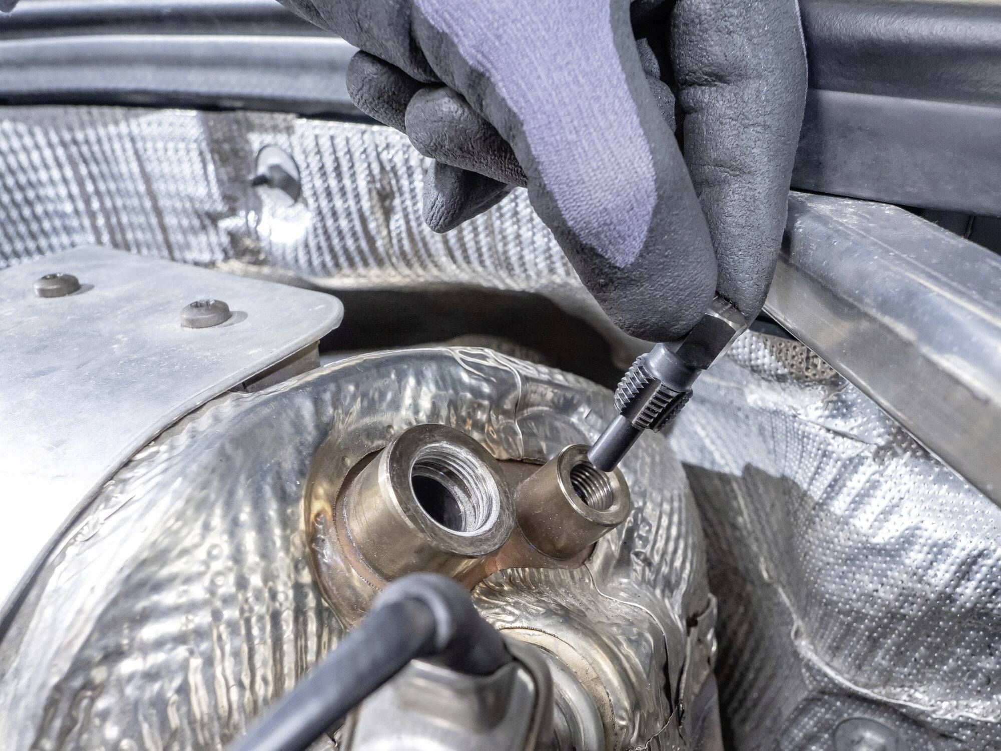 Eine Person mit Handschuhen repariert oder überprüft einen Anschluss an einem metallischen Autoteil unter der Motorhaube.