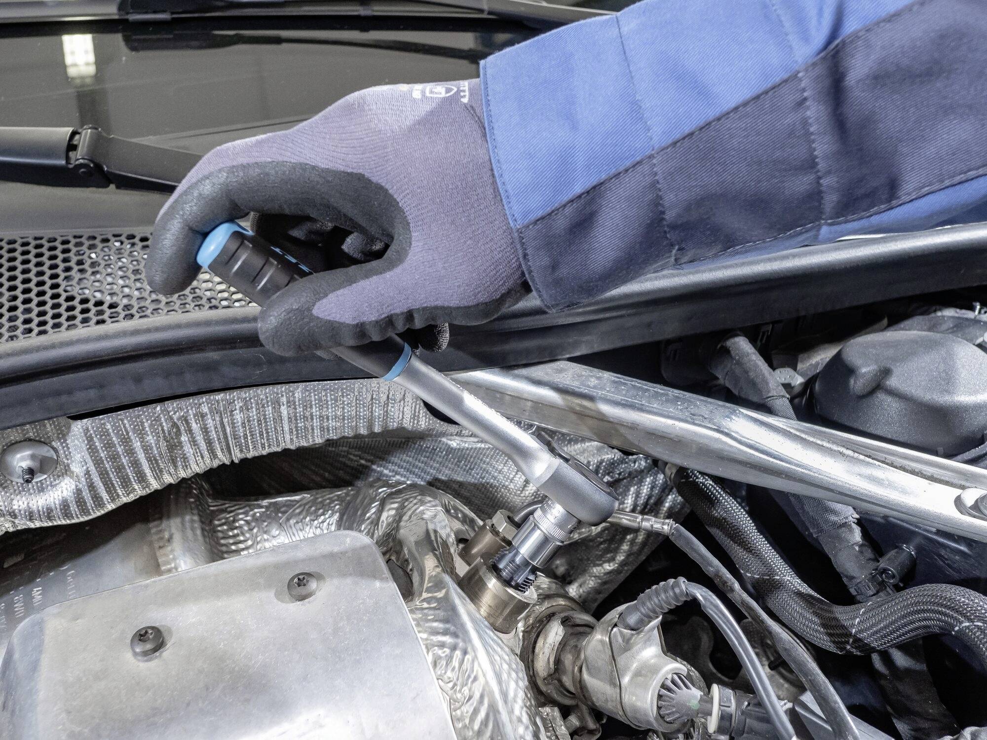Eine Hand in Handschuhen benutzt ein Werkzeug, um an einem Motor im Auto zu arbeiten. Das Bild zeigt eine Autoreparatur-Situation.