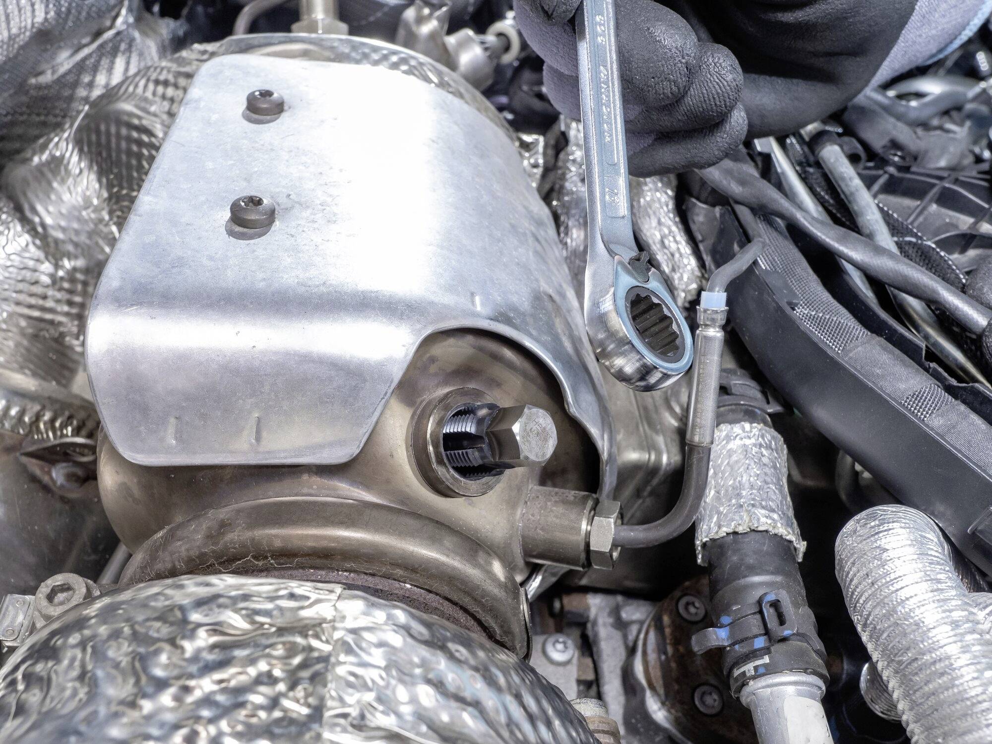 Mechaniker zieht mit einem Schraubenschlüssel eine Schraube im Motorraum eines Autos fest. Strukturierte Metallkomponenten sind sichtbar.