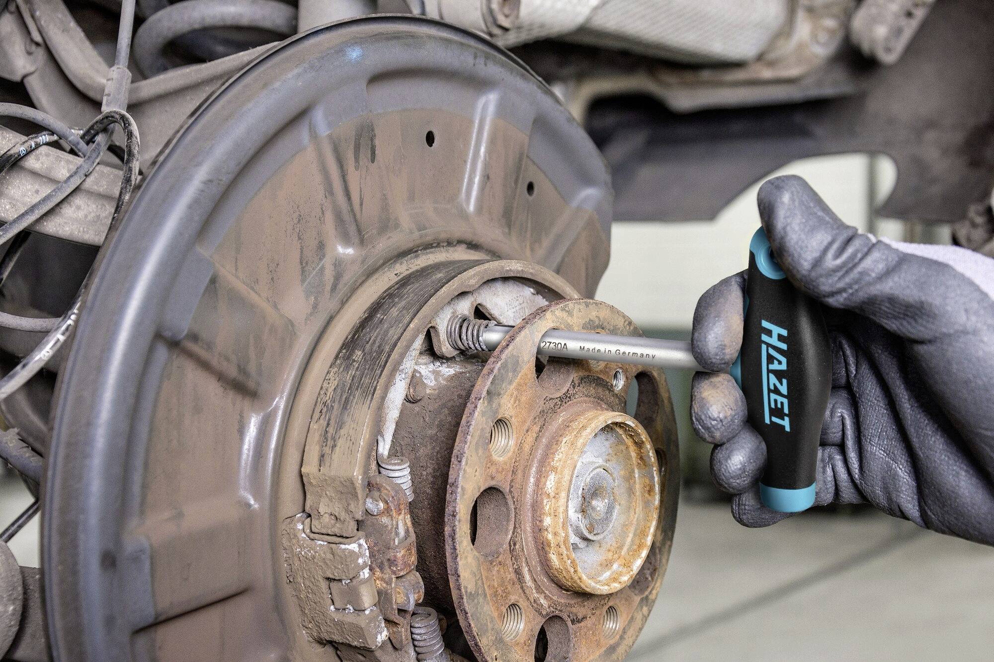 Ein Mechaniker zieht mit einem Werkzeug von Hazet eine Schraube an der Bremsanlage eines Autos fest. Fokus liegt auf der Bremsscheibe.
