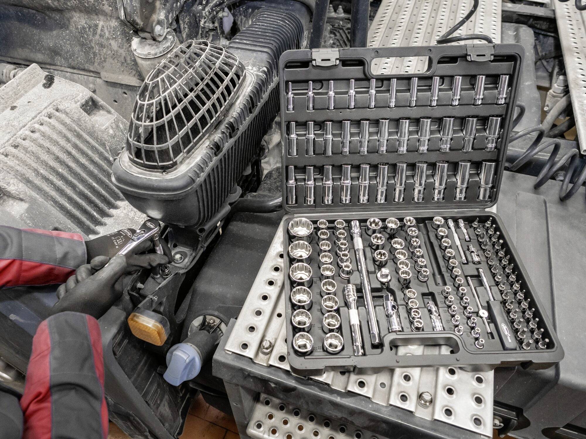 Mechaniker arbeitet mit einer Ratsche neben einem geöffneten Steckschlüsselsatz in einem Koffer auf einem Werkstatttisch.