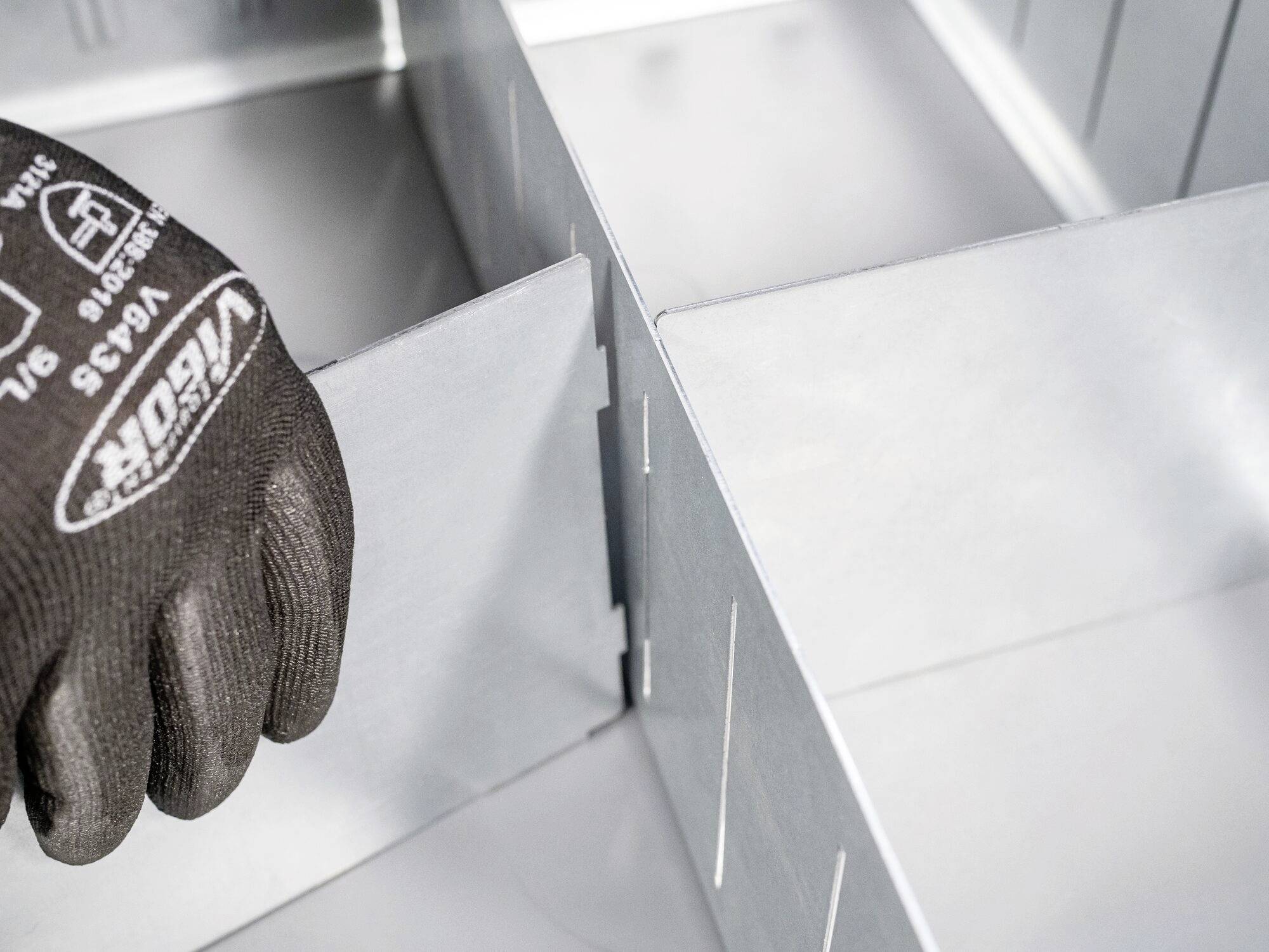 Eine Person mit einem schwarzen Handschuh fügt zwei silberne Metallplatten zusammen, vermutlich im Rahmen von Bauarbeiten oder Montage.