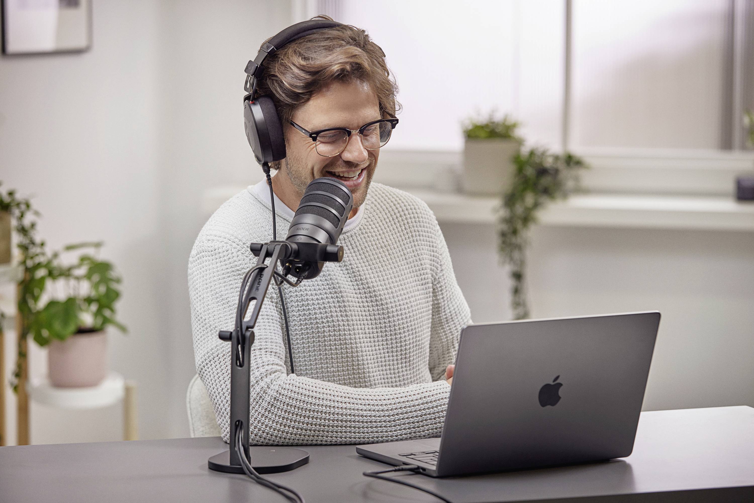 Ein Mann mit Brille und Kopfhörern sitzt lächelnd vor einem Mikrofon und einem Laptop in einem modernen, gut beleuchteten Raum.