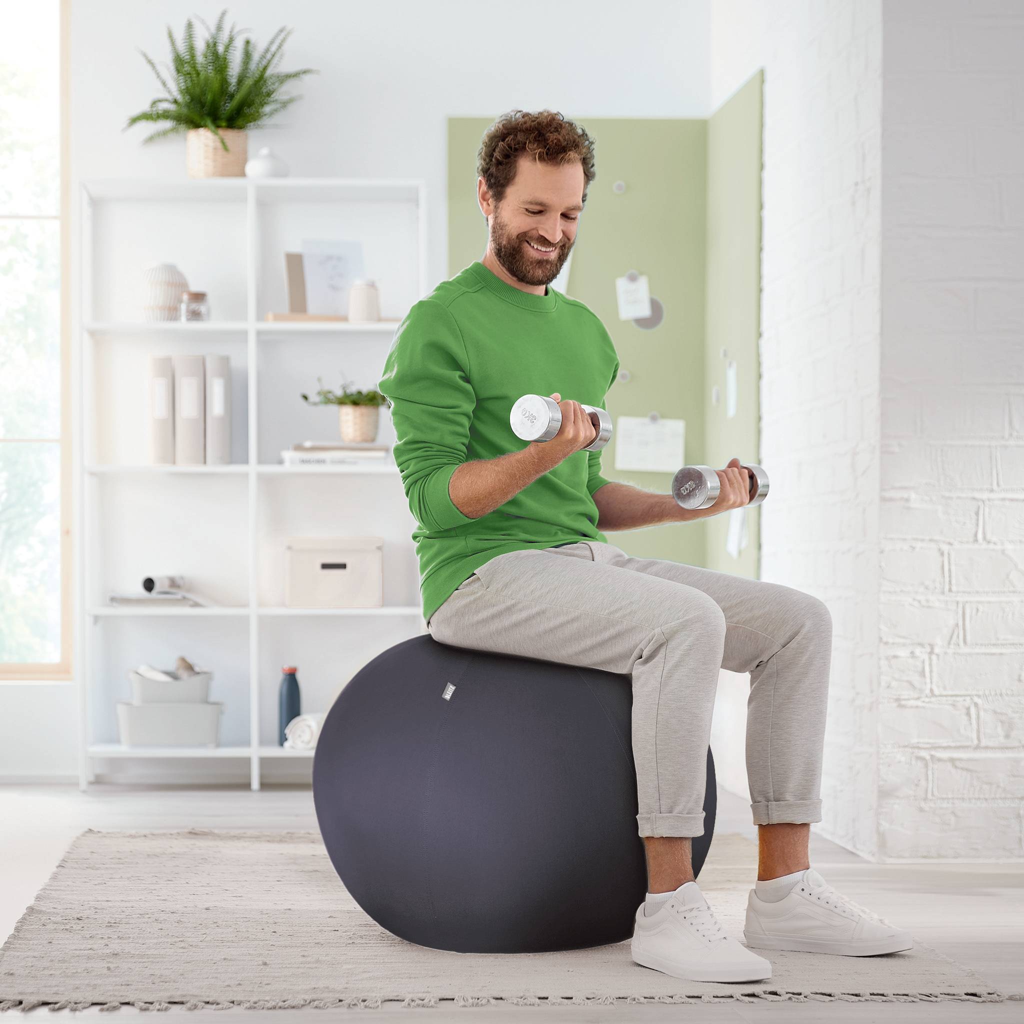Ein Mann trainiert mit Hanteln auf einem Gymnastikball in einem hellen Raum, umgeben von Pflanzen und Regalen.