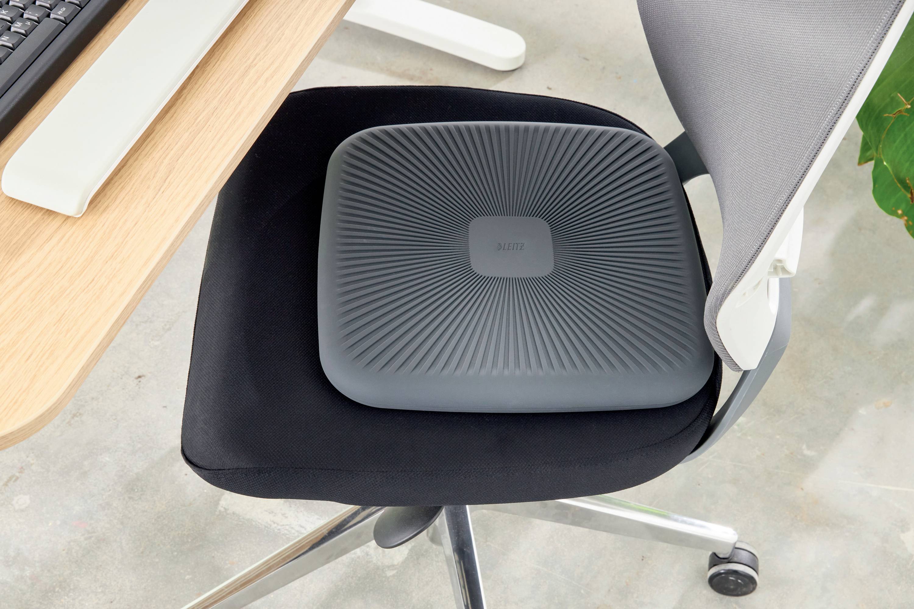 Sitzkissen auf einem Bürostuhl neben einem Schreibtisch mit Tastatur. Kissen ist dunkelgrau und quadratisch mit geriffelter Oberfläche.