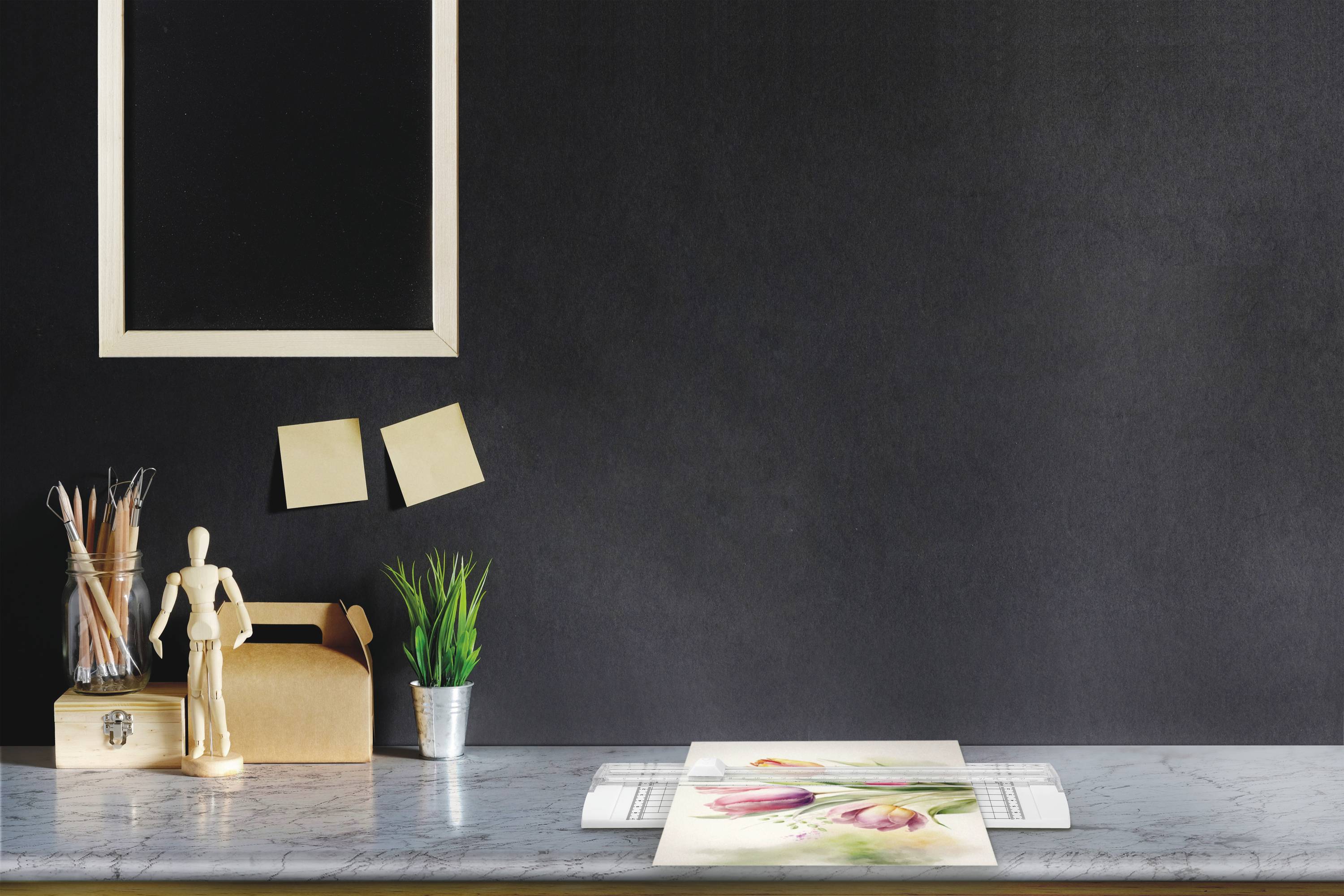 Ein Schreibtisch mit Zeichenutensilien, einem Notizblock und Pflanzen. An der Wand hängen zwei Notizzettel und ein leerer Bilderrahmen.