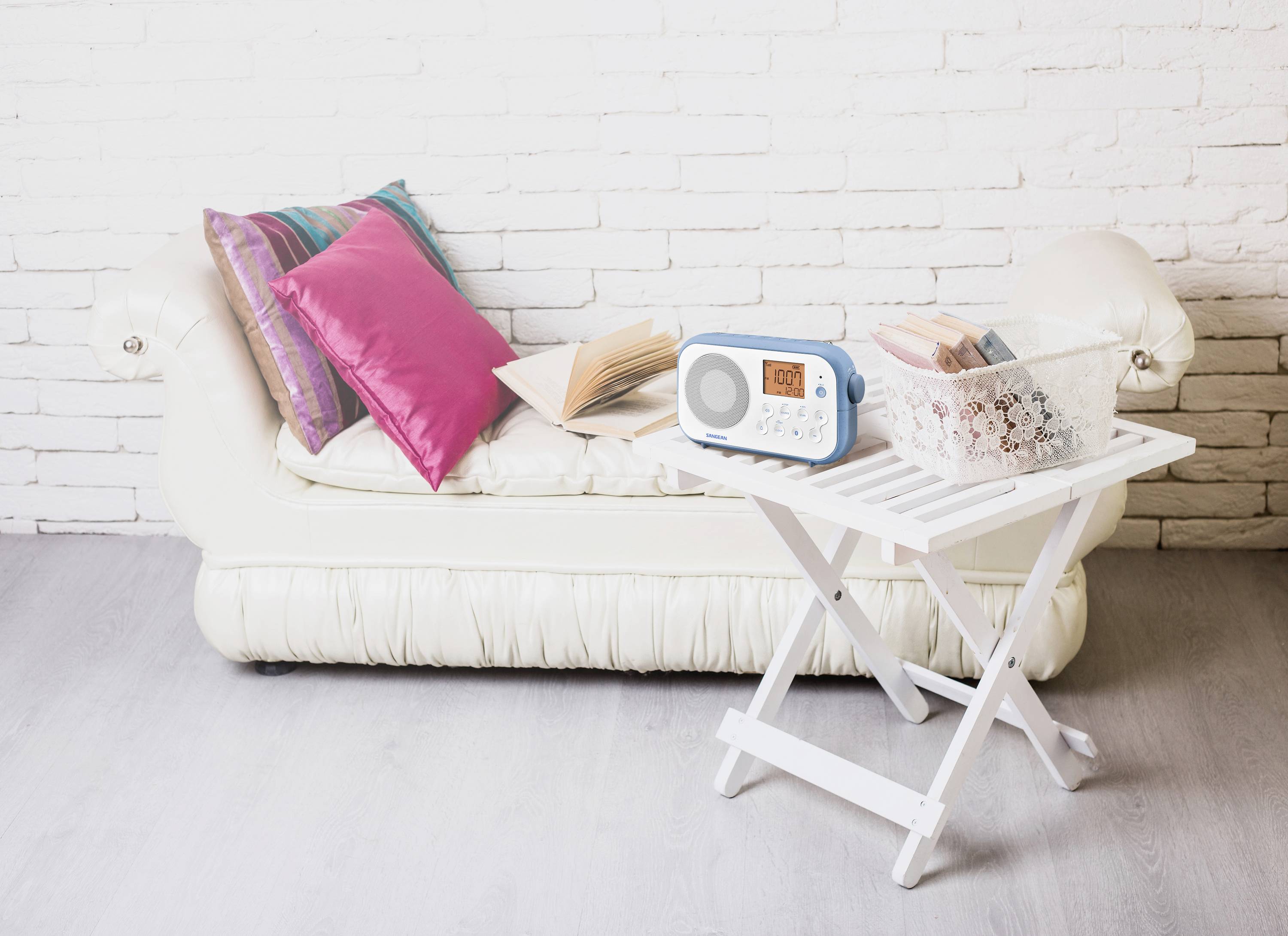 Ein weißes Sofa mit bunten Kissen steht vor einer weißen Backsteinwand. Ein offenes Buch liegt darauf. Ein weißer Tisch daneben hält ein Radio und einen Korb mit Fächern.