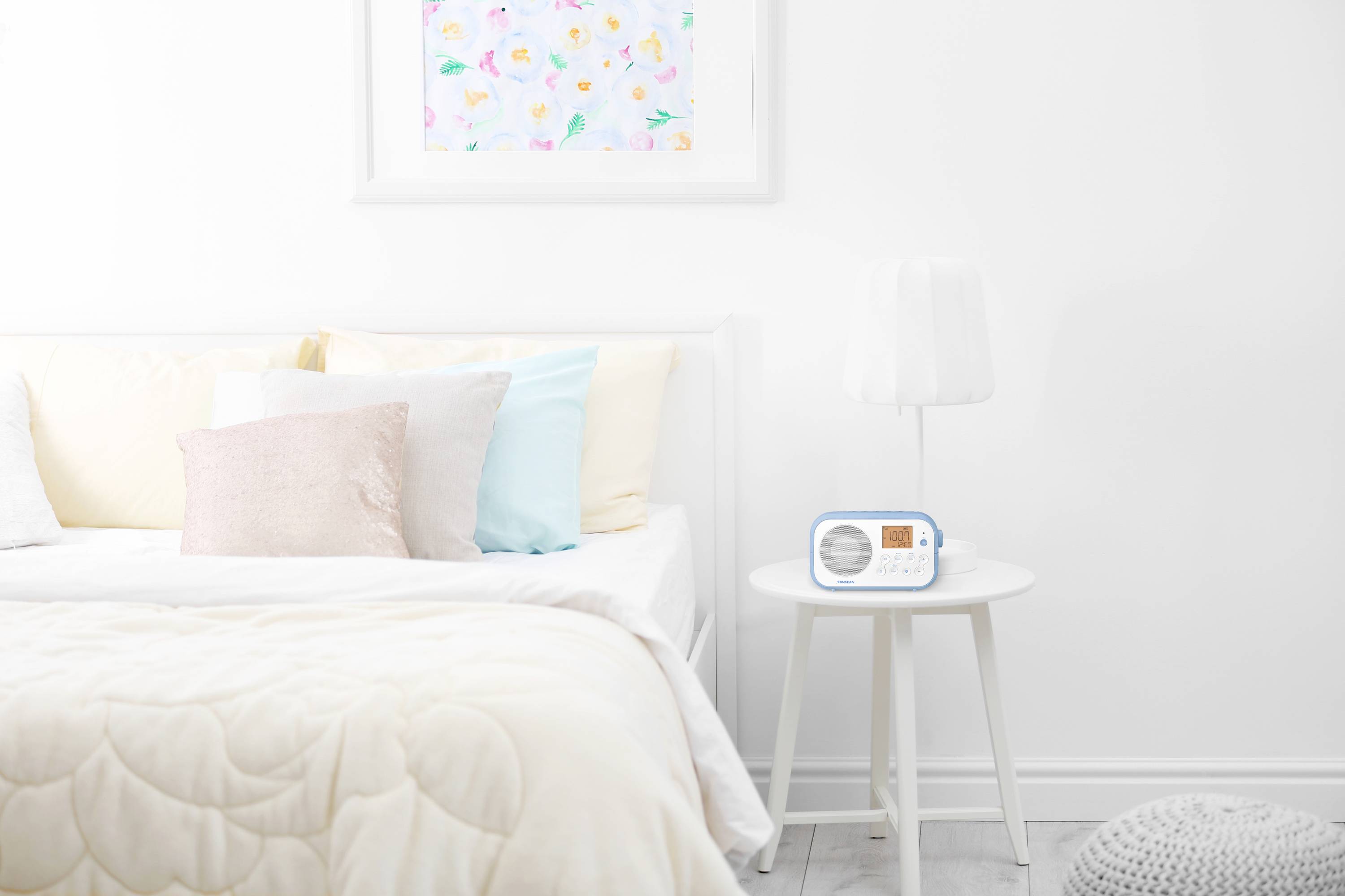 Helles Schlafzimmer mit weißem Bett, bunten Kissen und einem kleinen Tisch, auf dem ein weißes Radio steht. Buntes Bild an der Wand.