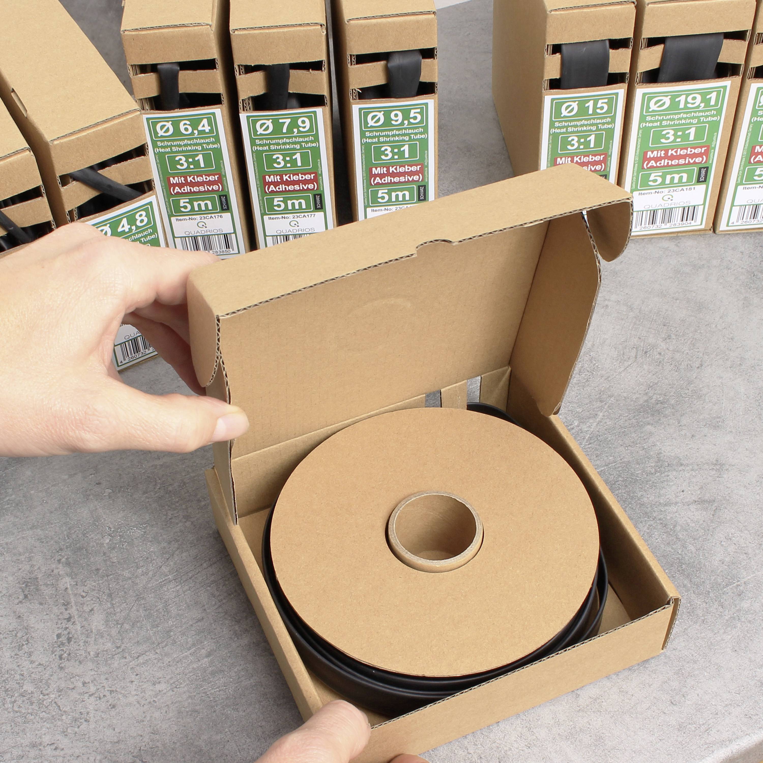 Person öffnet quadratische Kartonverpackung mit rundem schwarzen Klebeband. Weitere Kartons im Hintergrund stapeln sich.