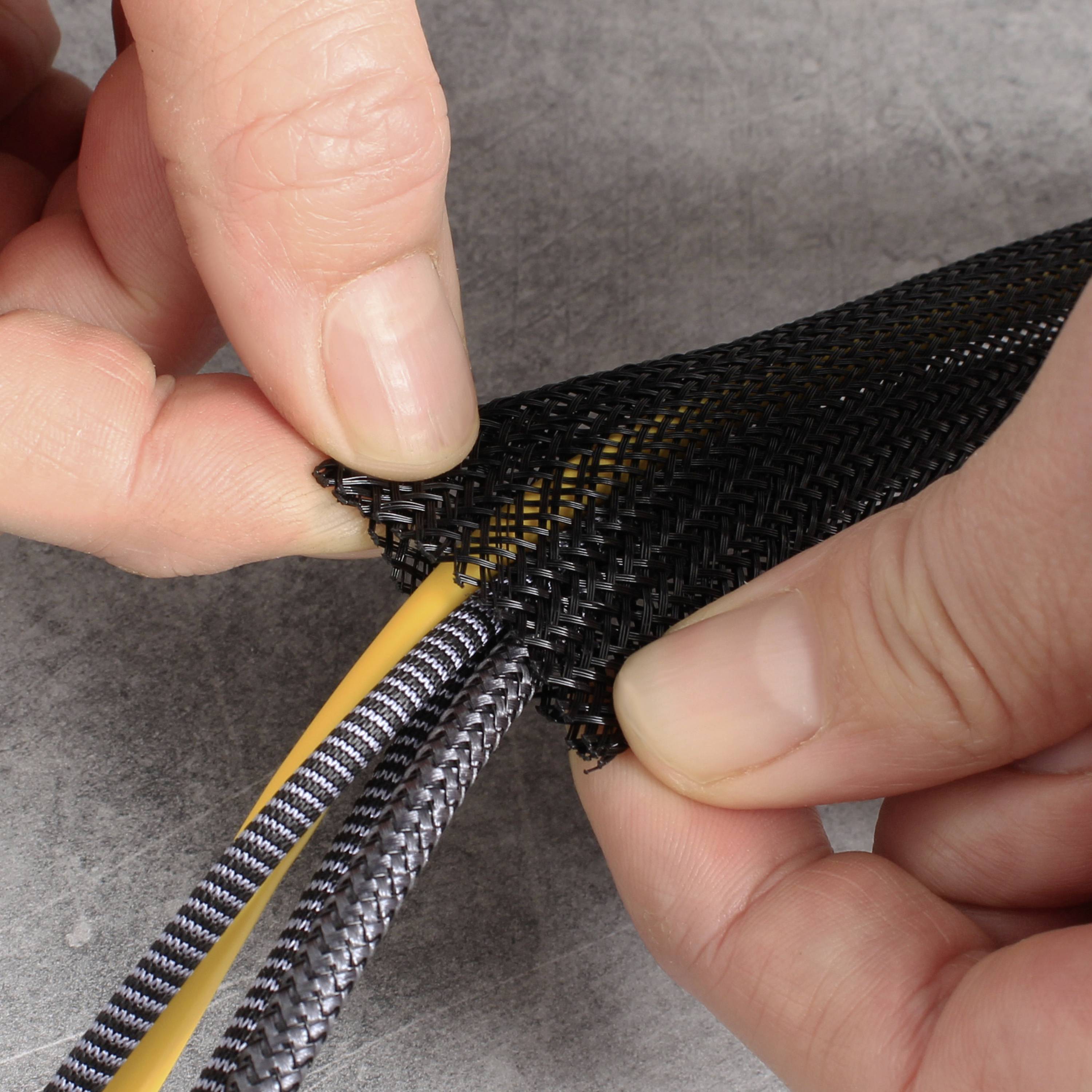 Hände flechten gelbe und schwarze Kabel in eine schwarz-graue Hülle. Nahaufnahme, Fokus auf Handarbeit und Textilstruktur.
