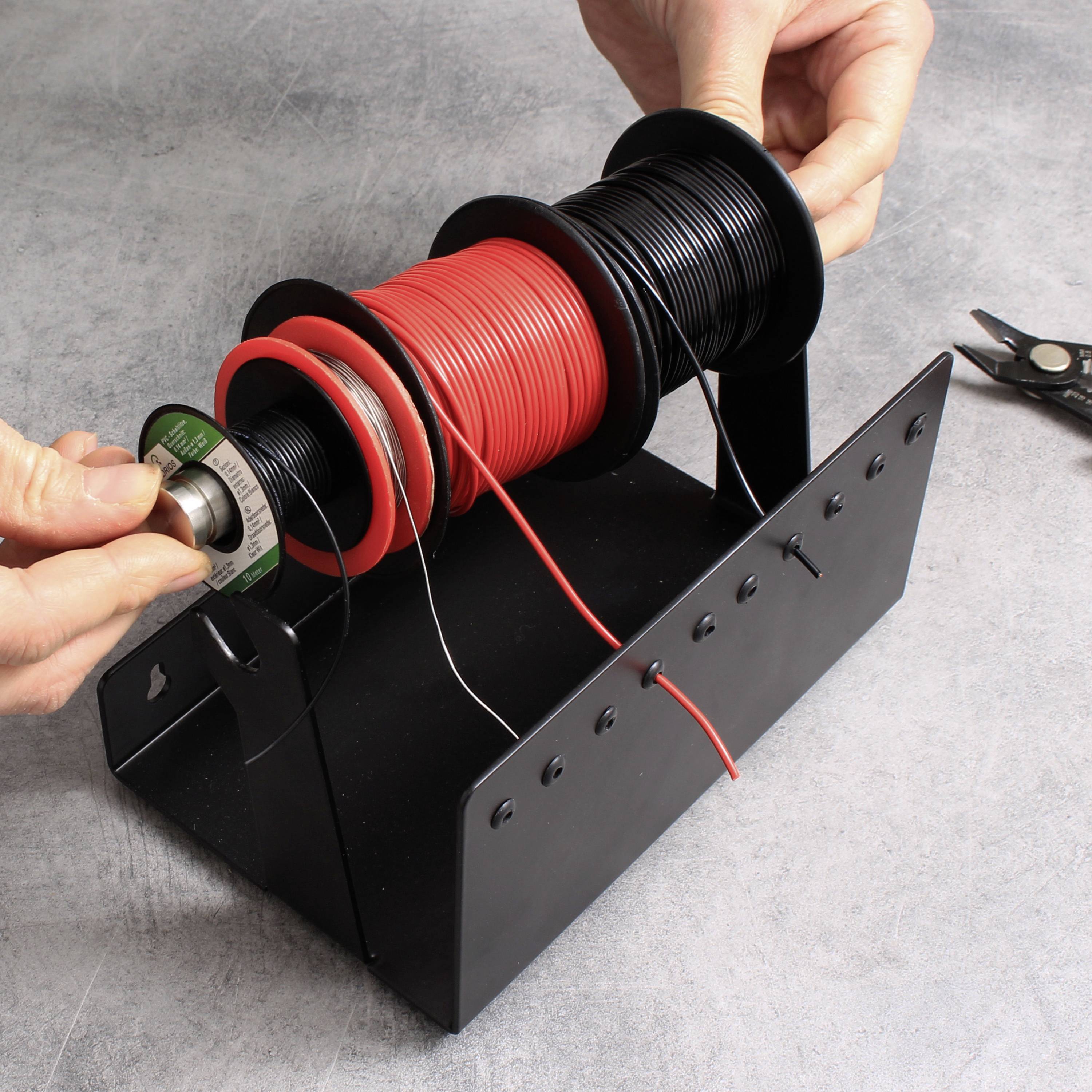 Szene zeigt Hände, die rote und schwarze Kabel von Spulen auf einem Metallhalter abrollen. Schneidewerkzeug liegt daneben.