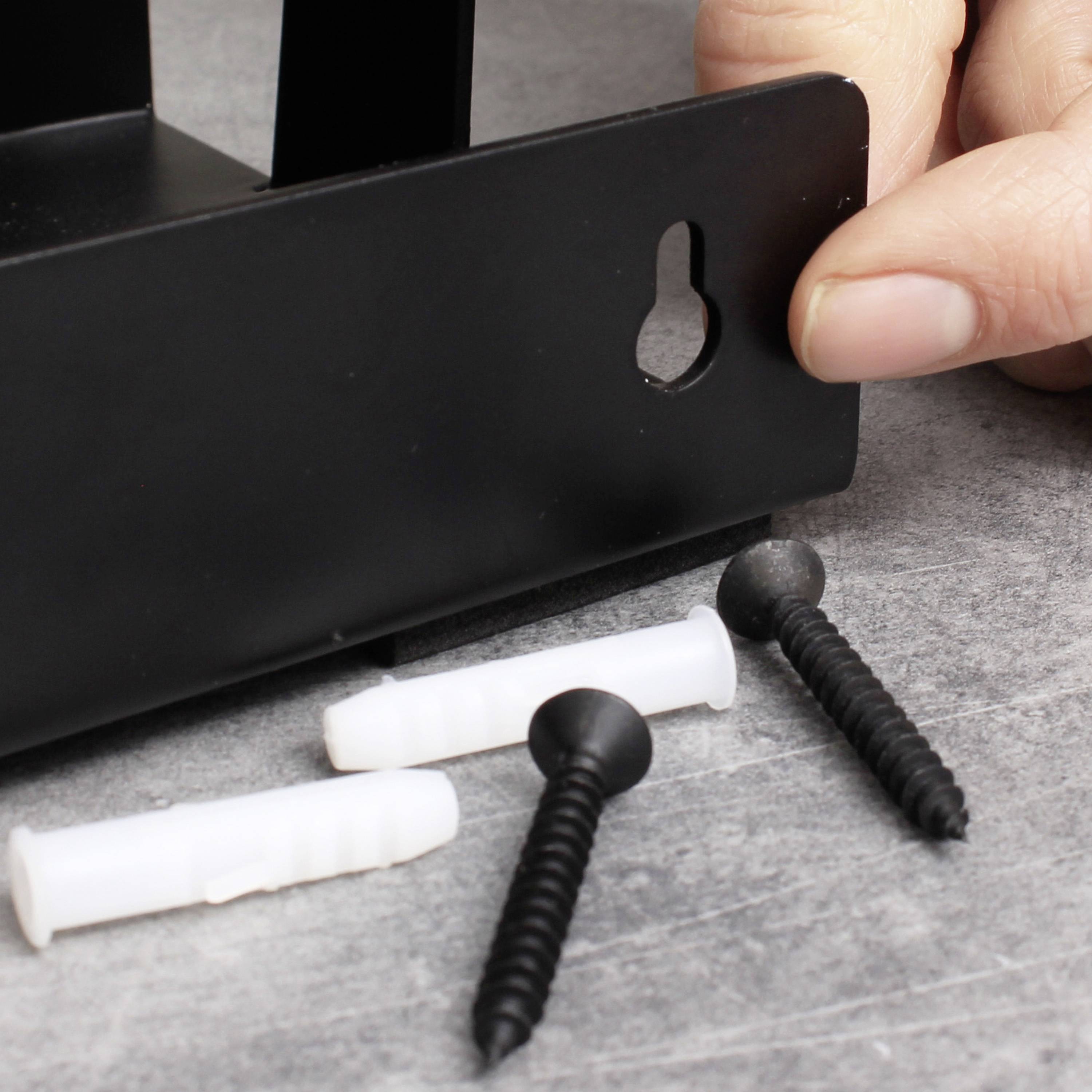 Hand hält ein schwarzes Metallteil mit Schlüssellochöffnung. Darunter liegen zwei Dübel und zwei Schrauben auf grauem Untergrund.