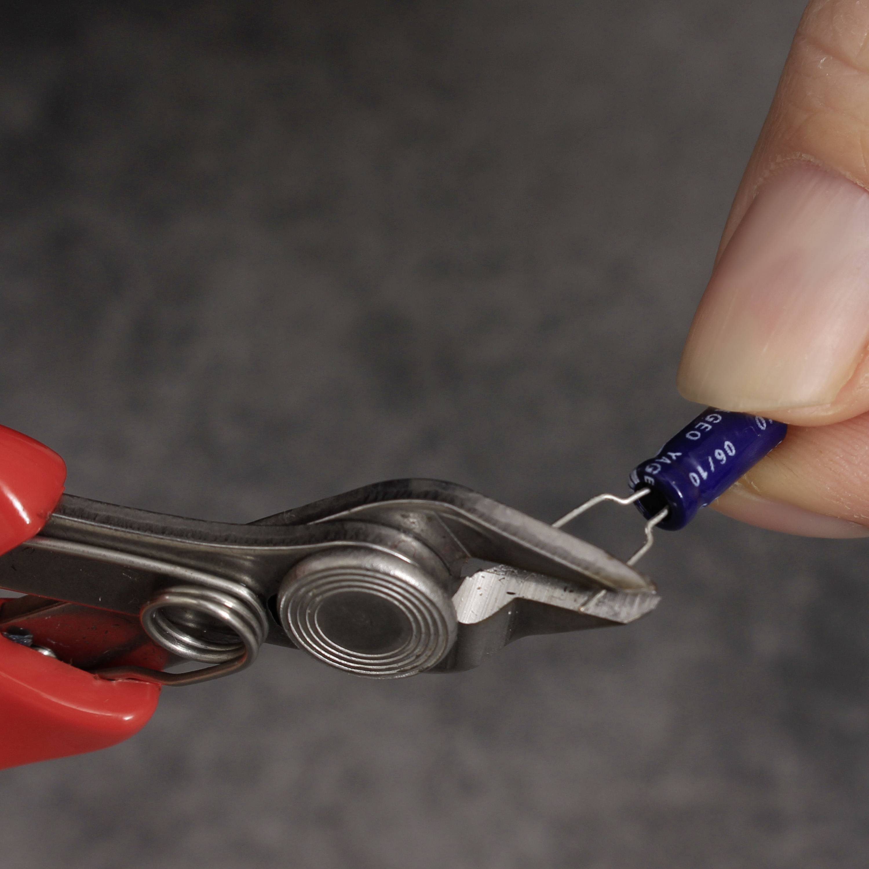 Eine Hand hält einen blauen Kondensator, der mit einer Krokodilklemme verbunden ist. Der Hintergrund ist verschwommen.