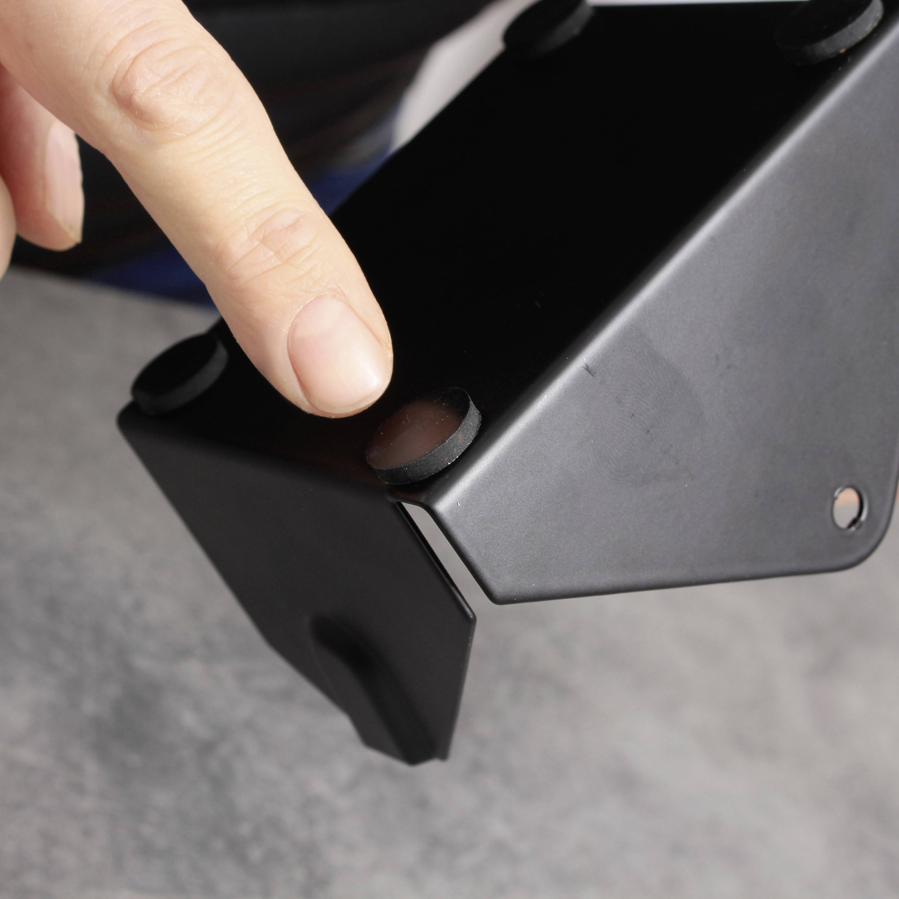 Ein Finger zeigt auf eine schwarze, rechteckige Halterung mit vier Gummifüßen, die auf einer grauen Oberfläche steht.