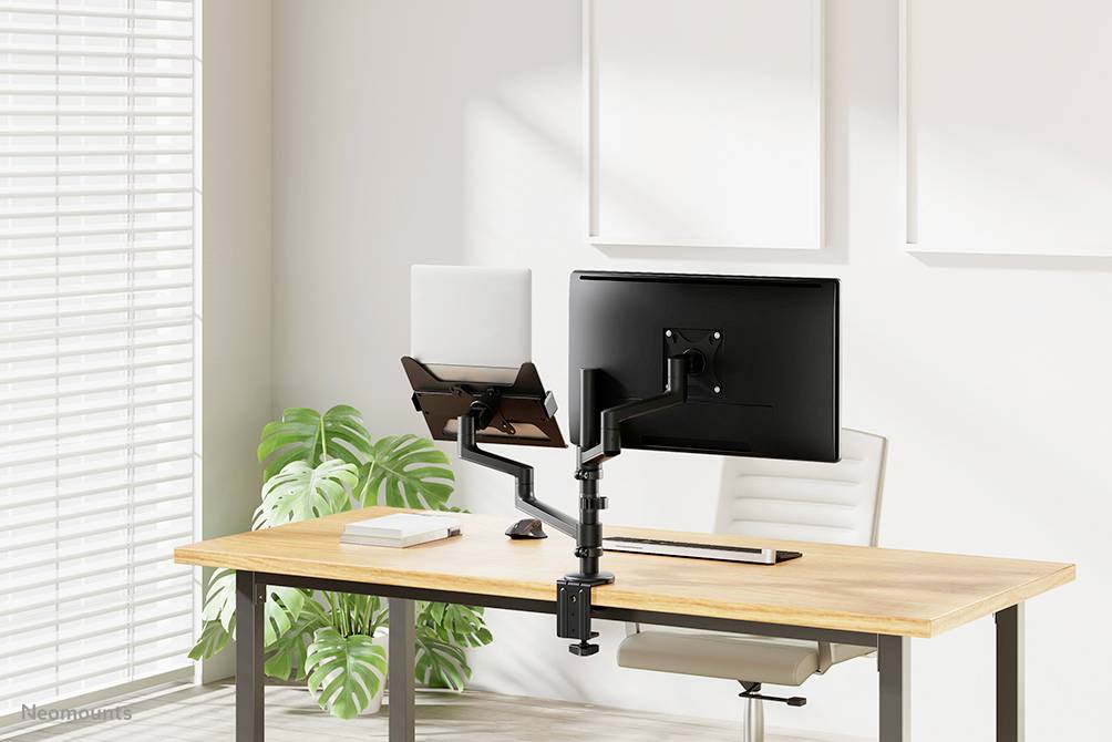 Ein moderner Schreibtisch mit Laptop und Monitor auf verstellbarem Halter, umgeben von natürlichem Licht und einer Pflanze.