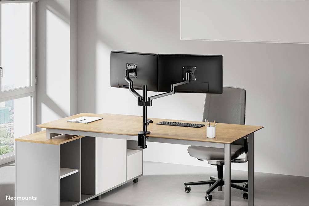 Zwei Monitore an einem Doppelschwenkarmhalter befestigt auf einem modernen Schreibtisch in einem hellen Büro mit Stuhl.