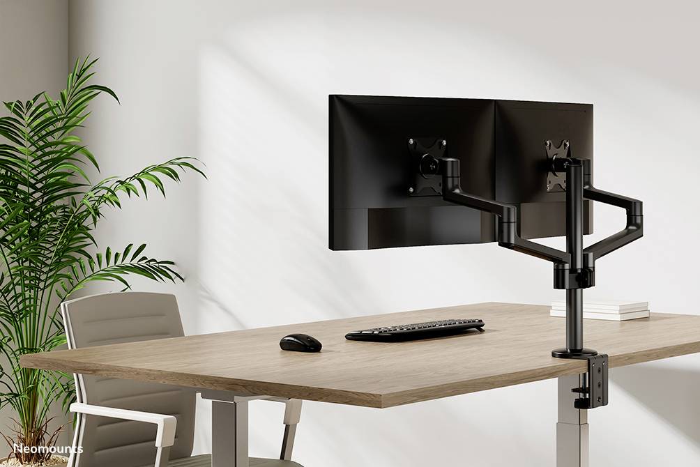 Zwei Monitore an einstellbaren Halterungen auf einem Schreibtisch mit Tastatur und Maus, daneben ein Bürostuhl und eine Pflanze.