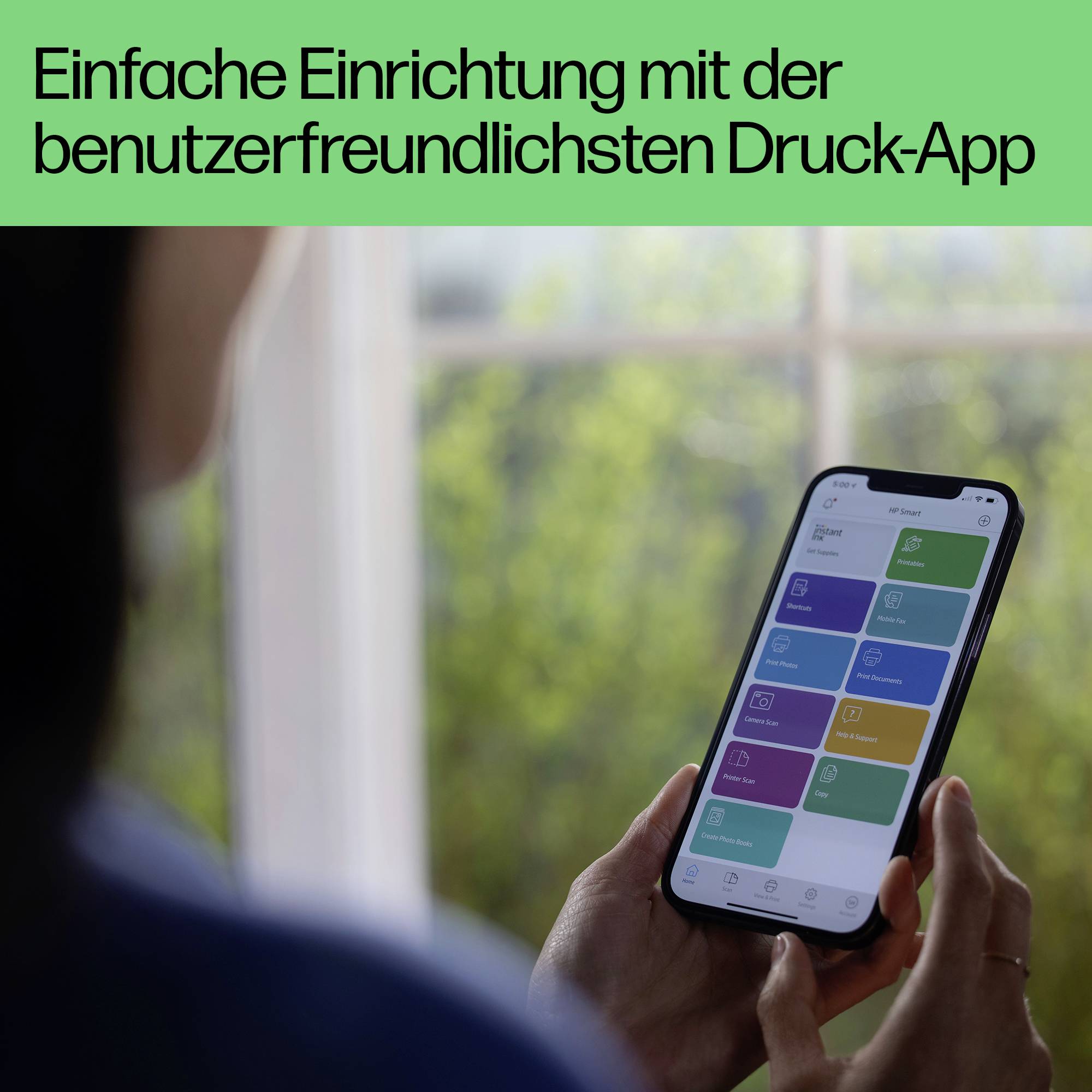 Person hält ein Smartphone mit geöffneter Druck-App; im Hintergrund ist ein Fenster erkennbar. Text: 'Einfache Einrichtung mit der benutzerfreundlichsten Druck-App'.