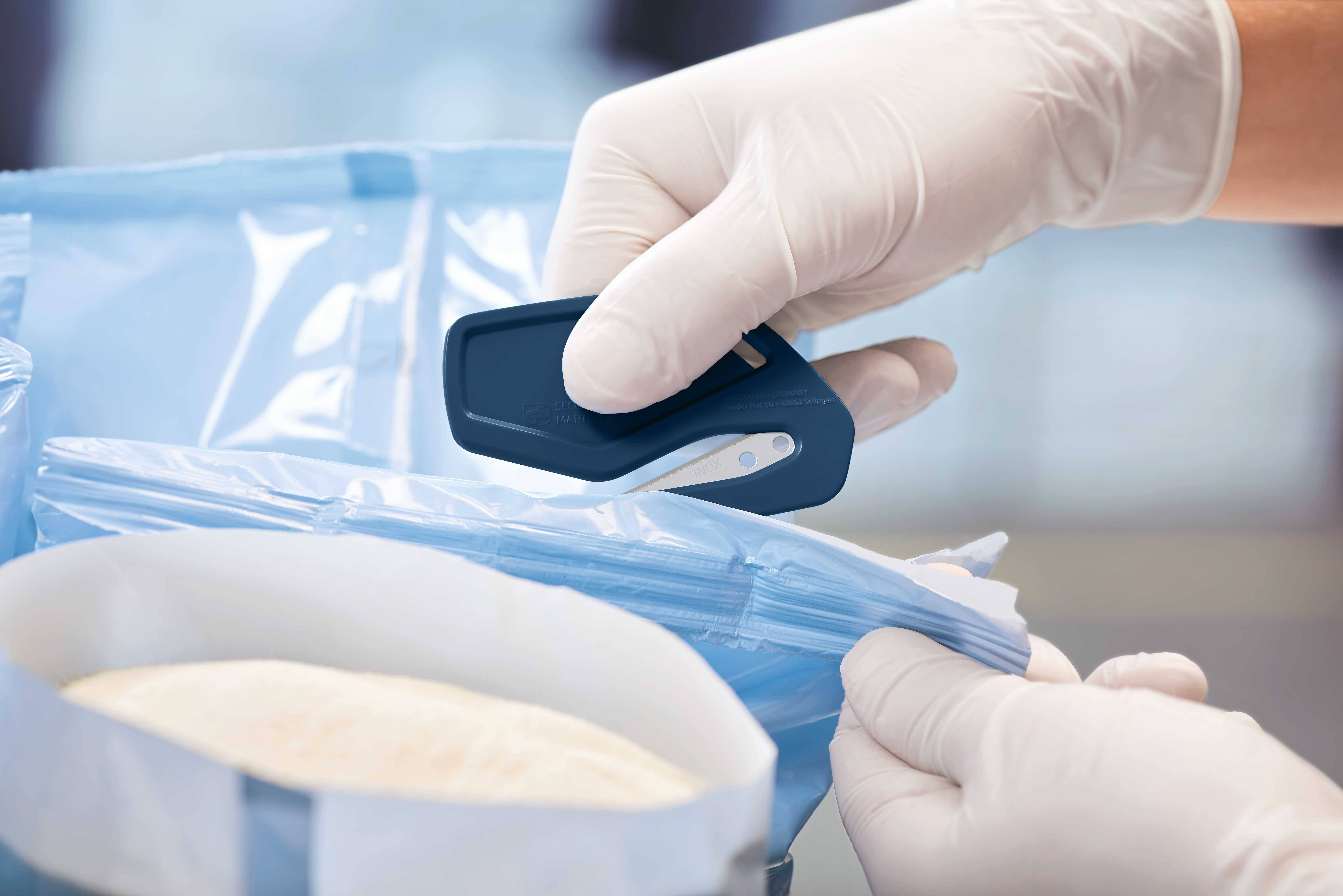 Eine Person öffnet mit einem blauen Verschlussaufschneider einen Plastikbeutel. Die Person trägt weiße, medizinische Handschuhe.