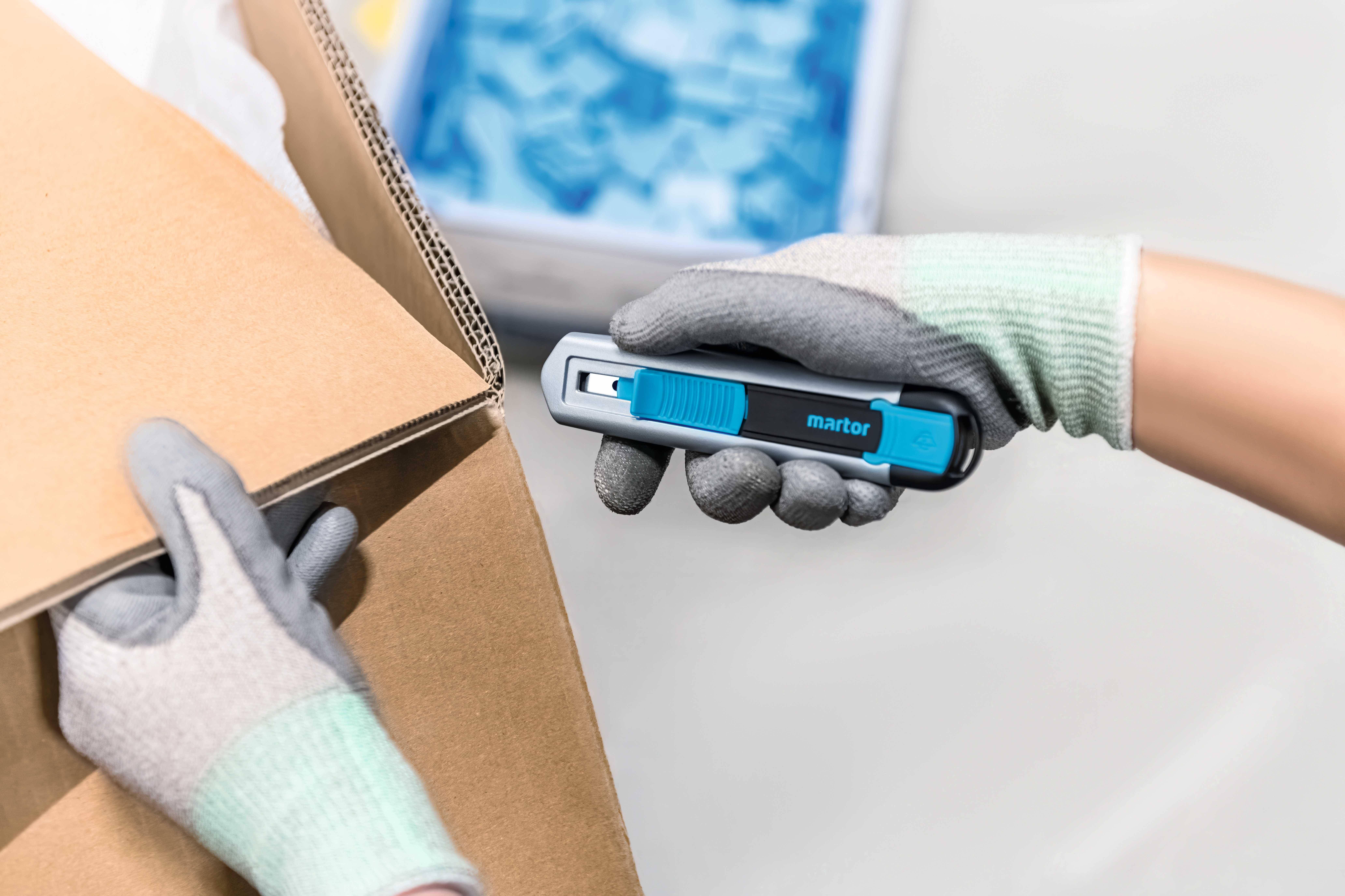 Eine Person mit Handschuhen schneidet mit einem blauen Sicherheitsmesser einen Karton auf. Im Hintergrund sind blaue Objekte zu sehen.