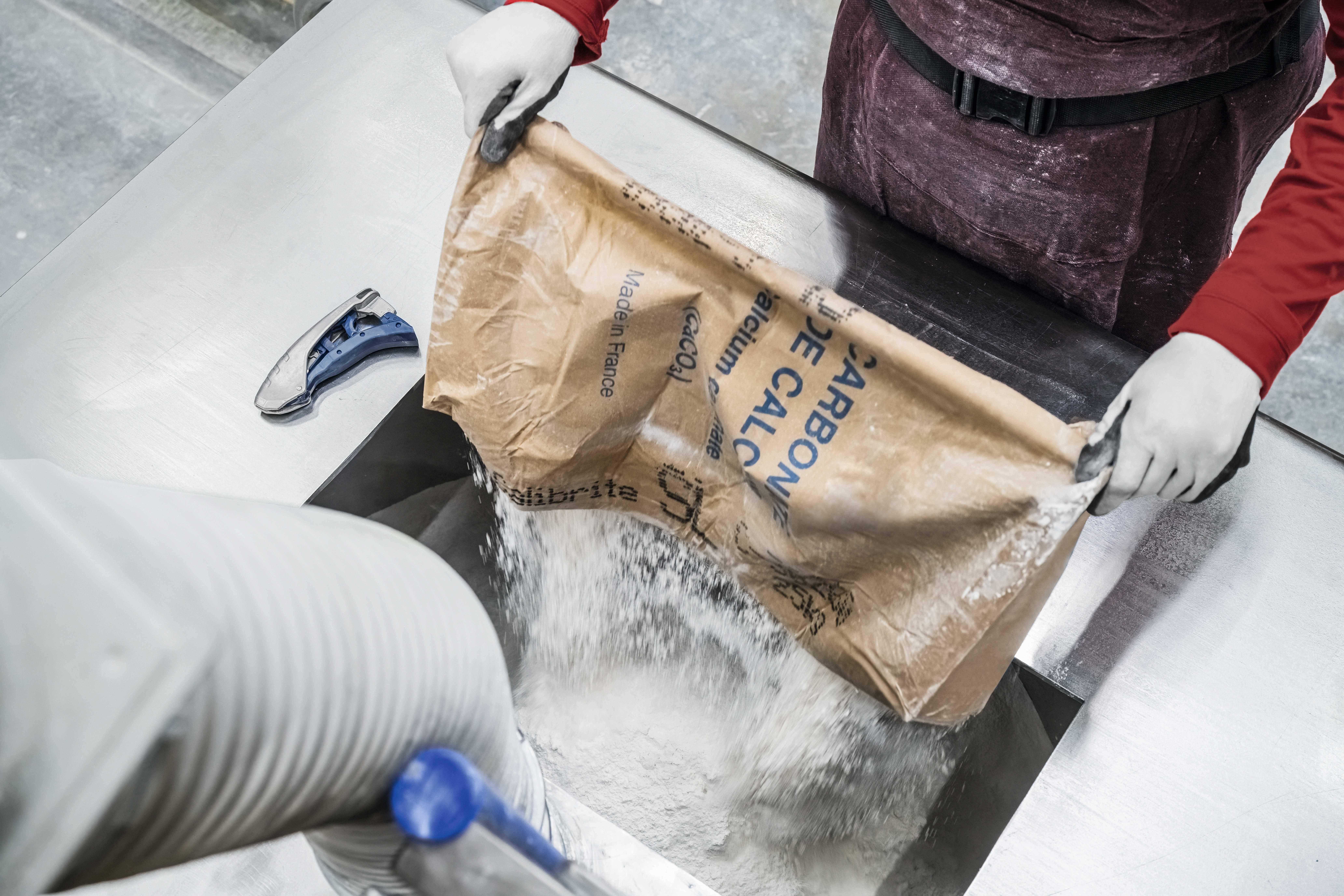 Eine Person in Handschuhen leert einen Papiersack mit Kalkpulver in eine industrielle Maschine. Ein Handgerät liegt daneben.