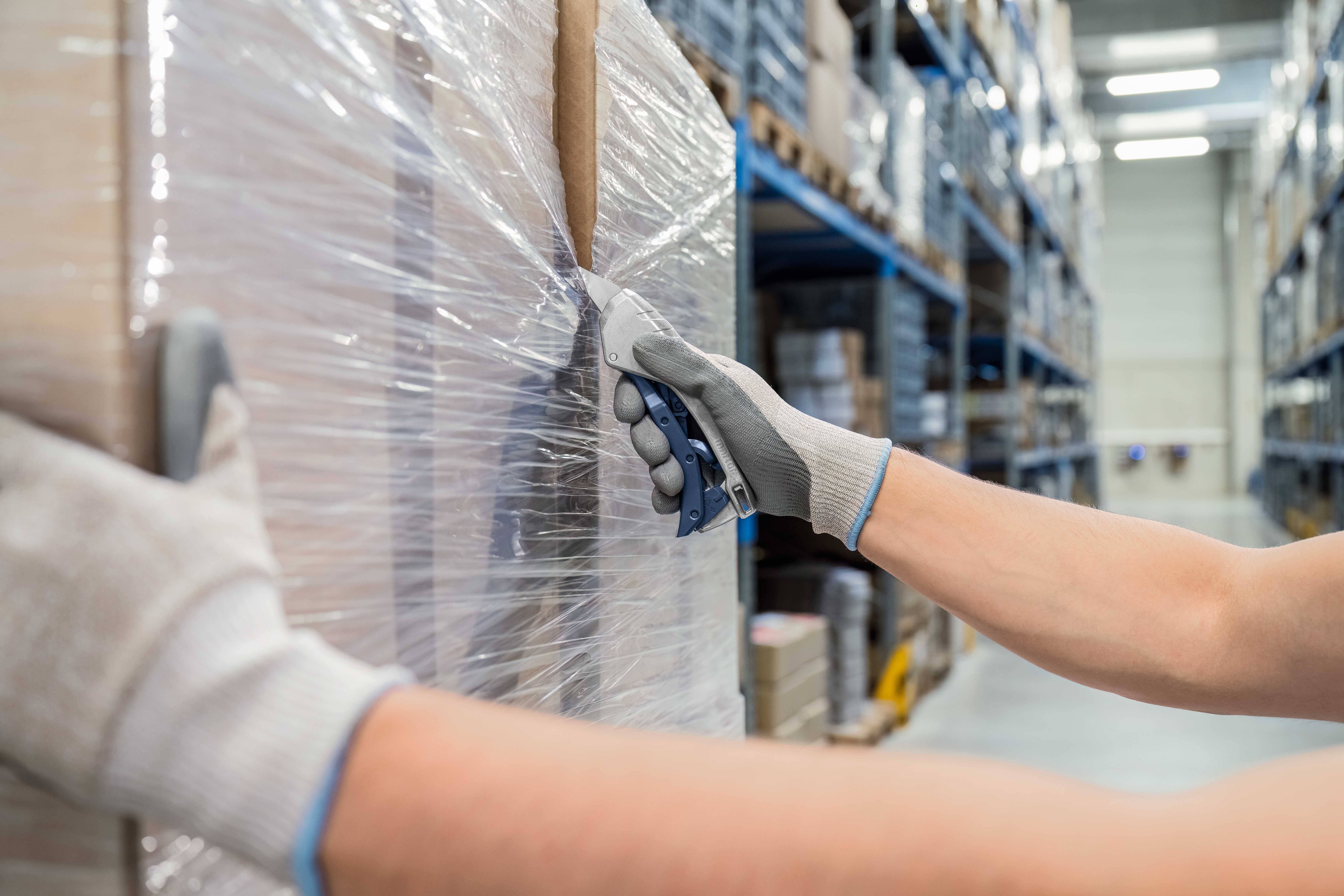 Person schneidet Plastikfolie von einer Palette in einem Lager ab, während sie Arbeitshandschuhe trägt. Regale voller Kartons im Hintergrund.