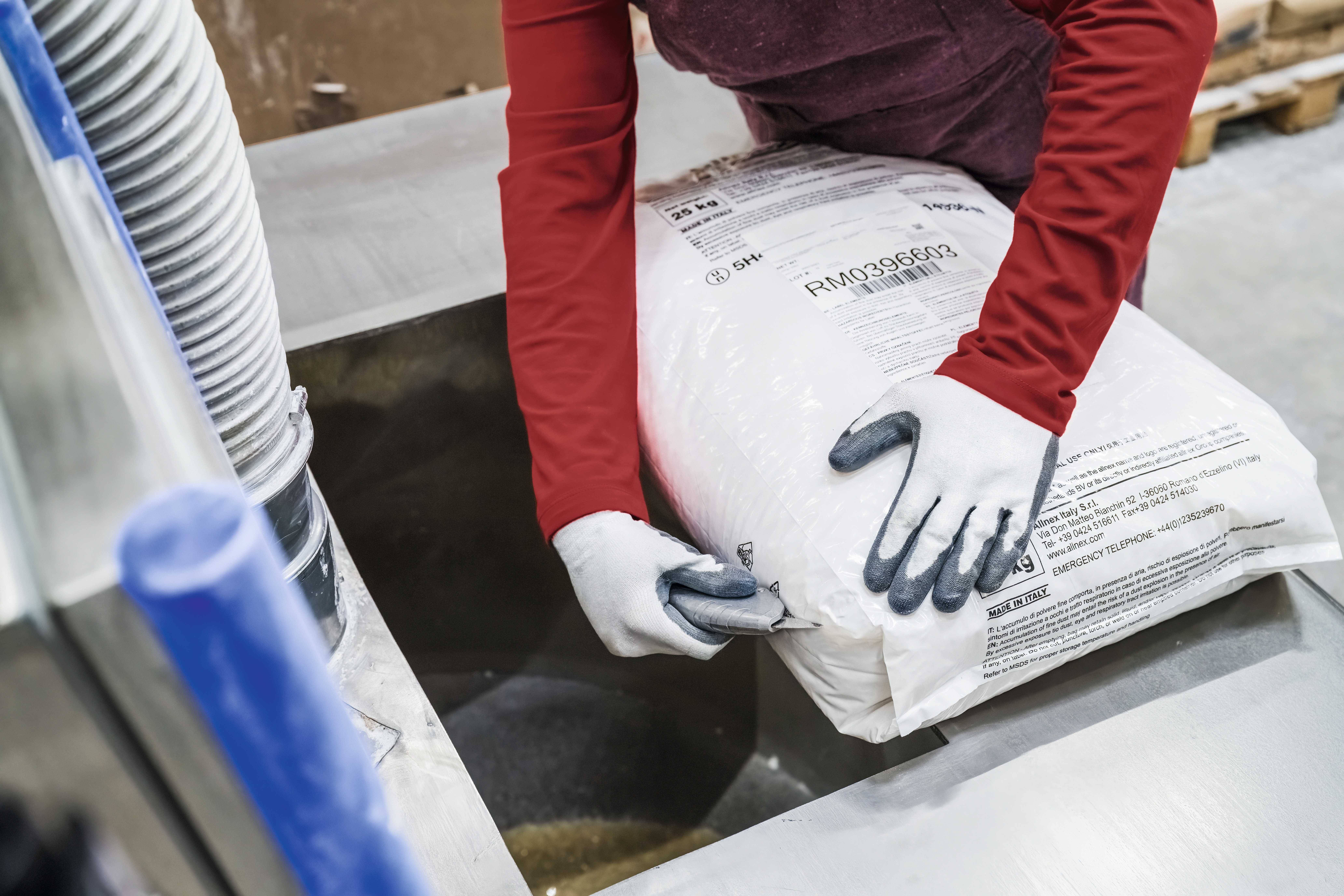 Eine Person in Handschuhen entleert einen Sack mit Material in einen Trichter in einer industriellen Umgebung.