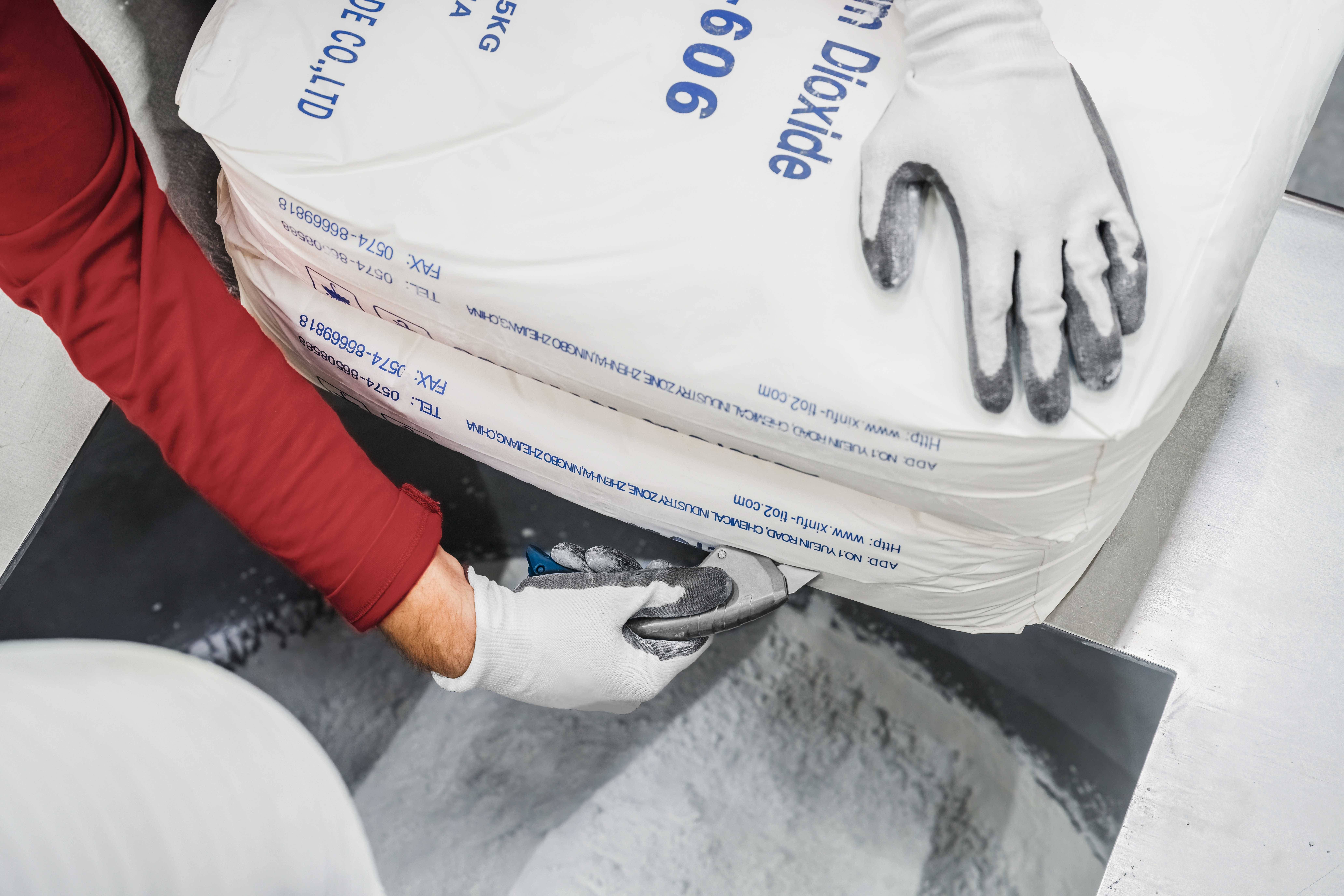 Eine Person in roten Ärmeln öffnet einen großen Sack mit weißem Pulver auf einer Waage. Die Hand trägt weiße Handschuhe.