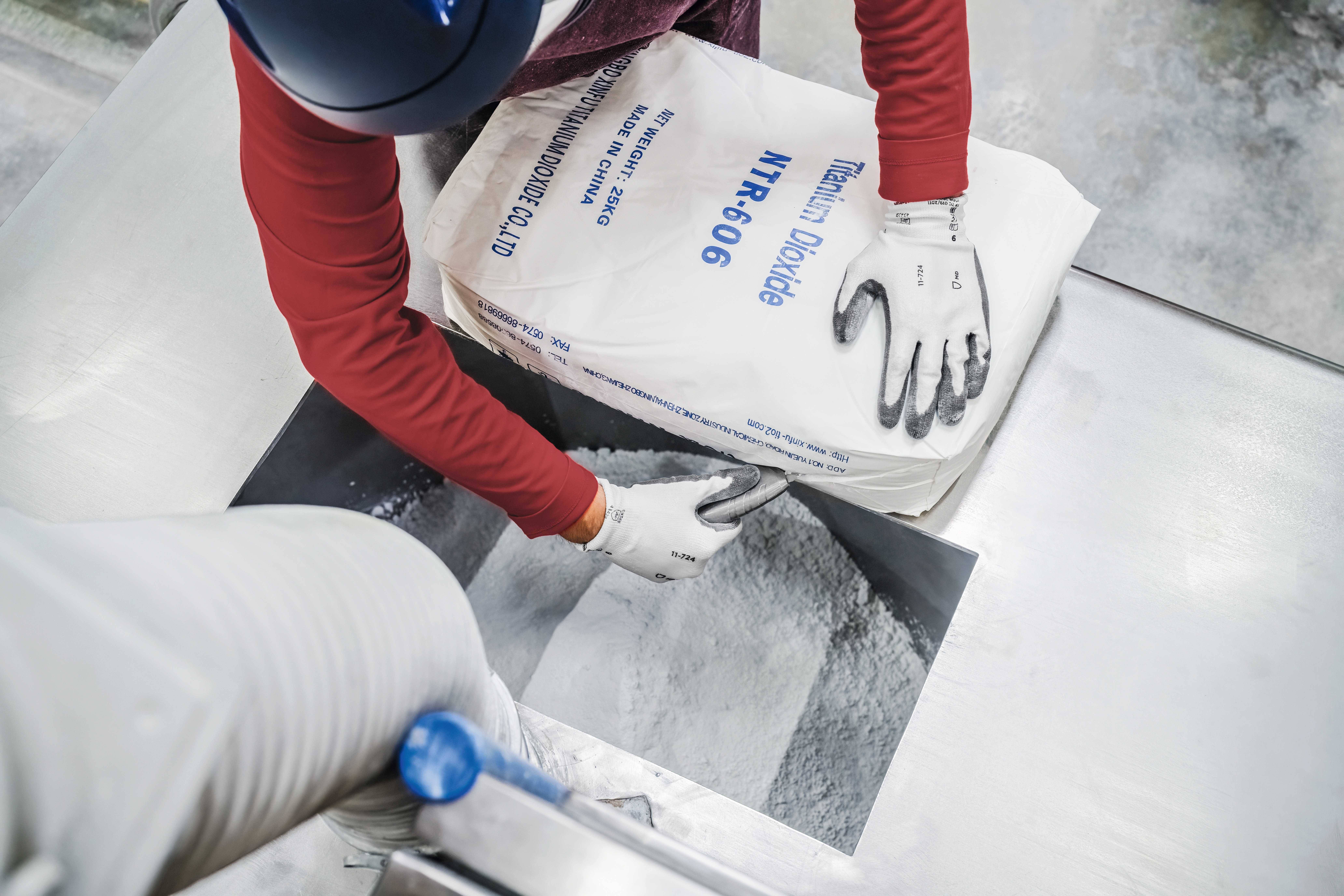 Eine Person mit Handschuhen gießt Titan-Dioxid aus einem beschrifteten Sack in eine Maschine. Industrialle Umgebung.