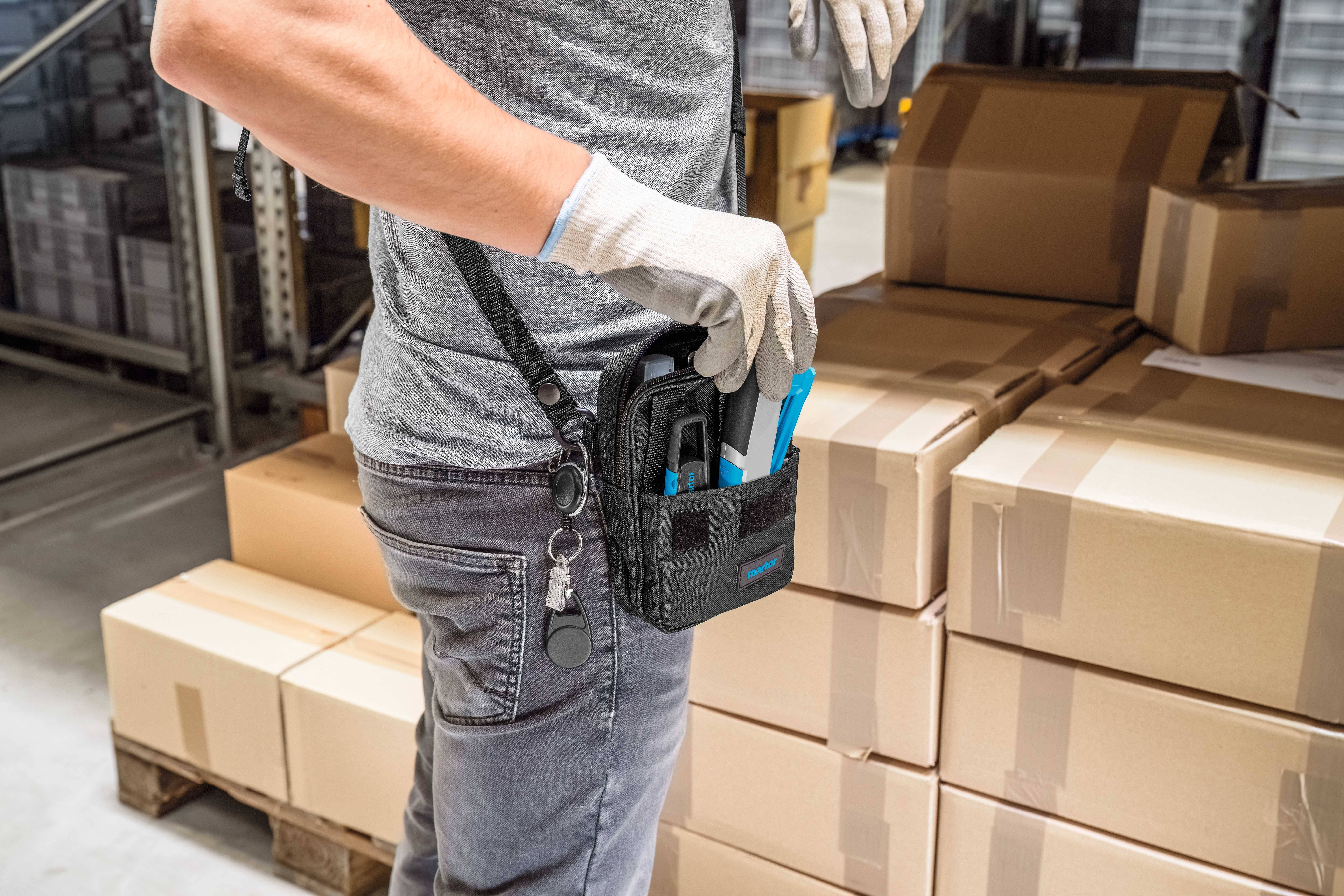 Person packt Werkzeuge aus einer Tasche am Gürtel neben gestapelten Kartons in einem Lager aus.