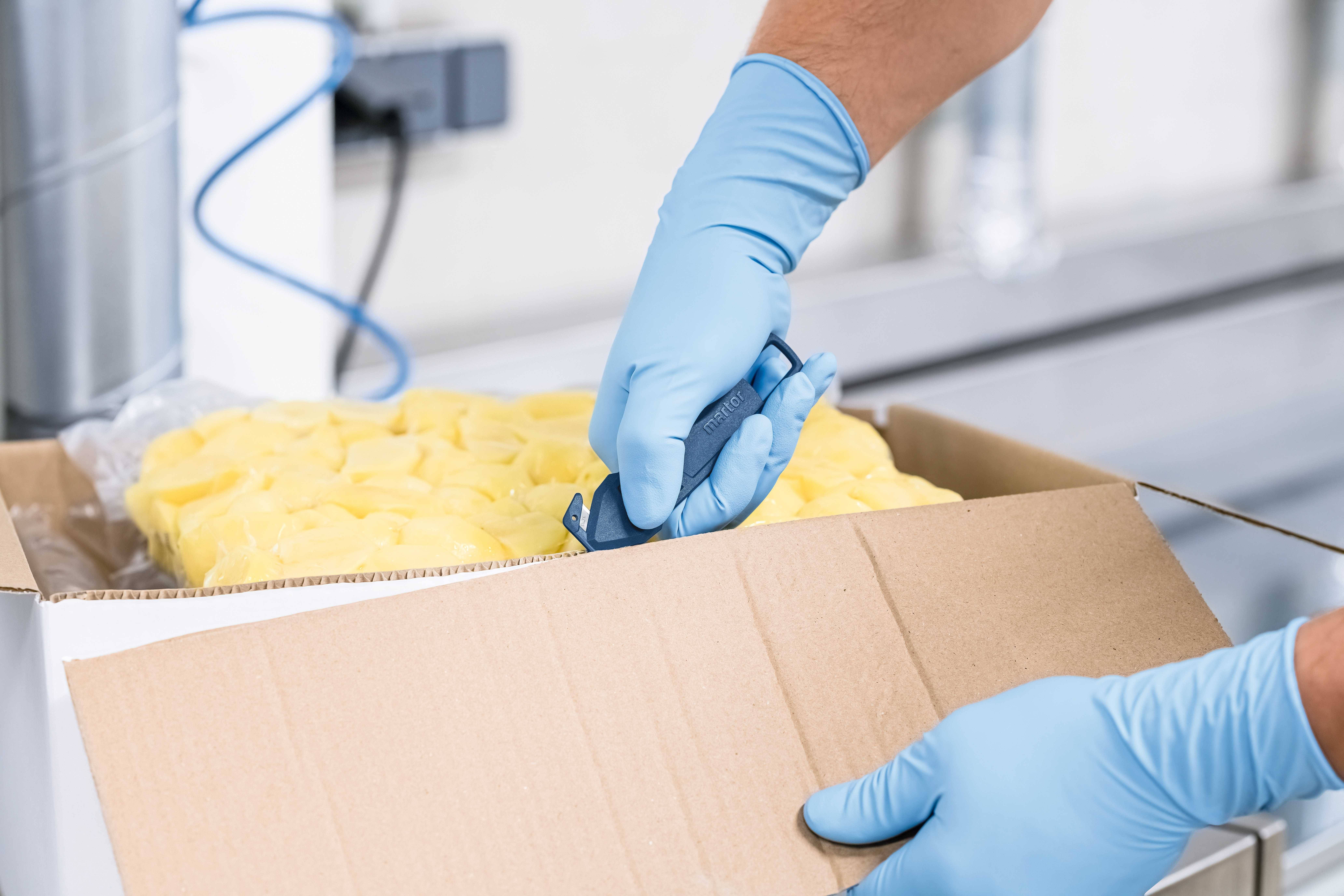 Person mit blauen Handschuhen öffnet mit einem Messer einen Karton mit verpackten gelben Objekten auf einem Tisch in einer Fabrikumgebung.