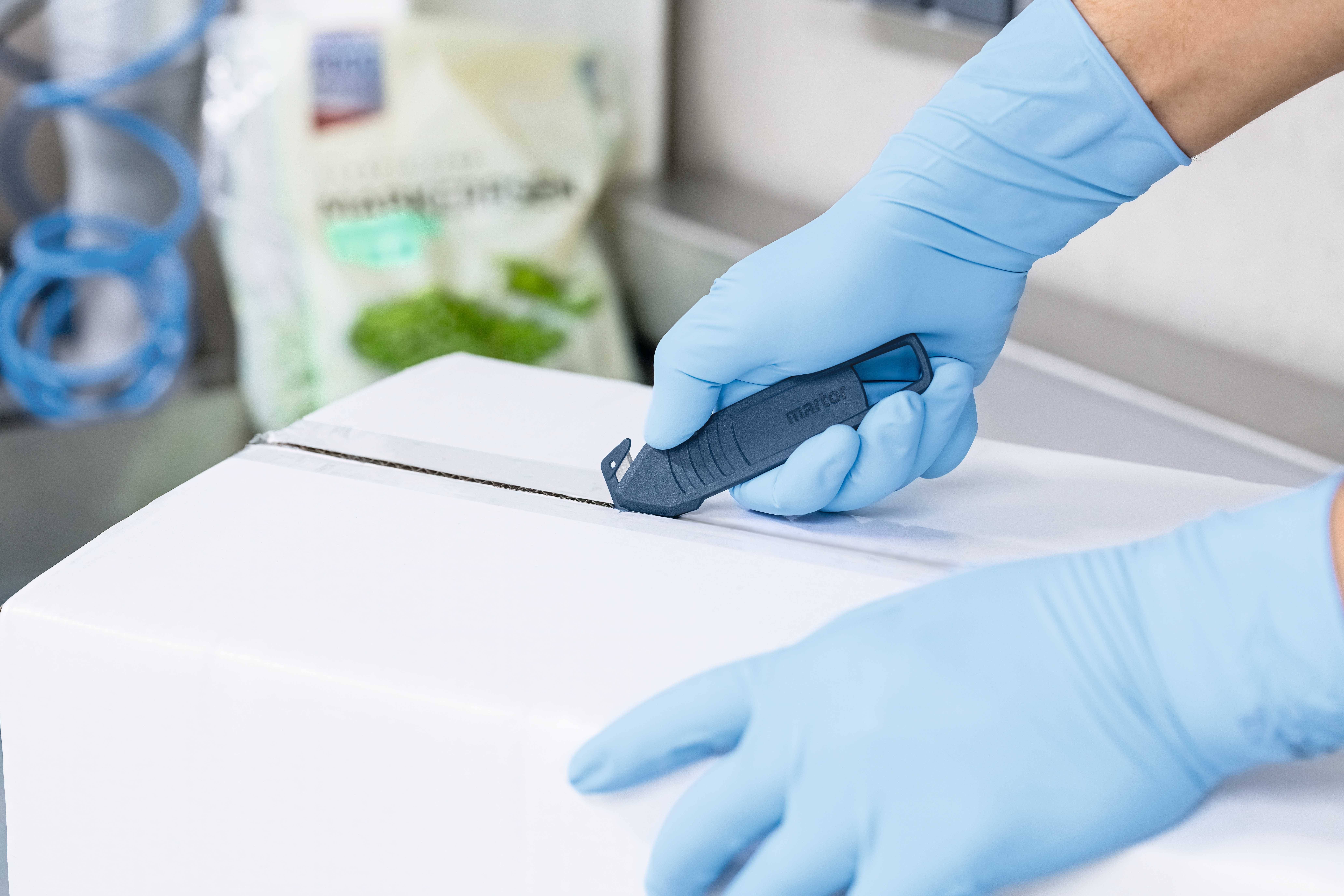 Eine Person schneidet mit einem Sicherheitsmesser einen Karton. Sie trägt blaue Einweghandschuhe. Im Hintergrund ist verschwommen eine Packung Salat zu sehen.