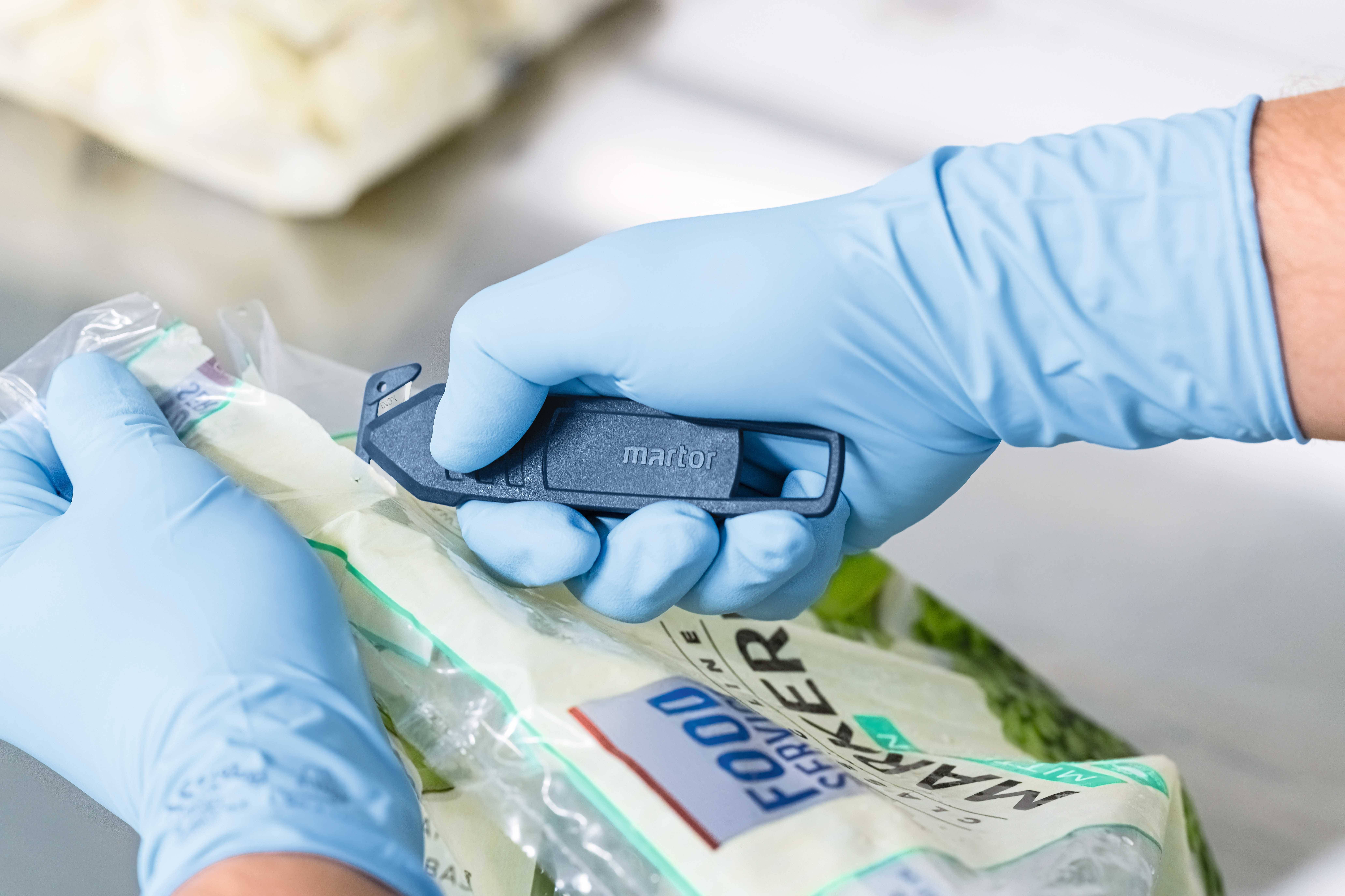 Person mit blauen Handschuhen öffnet eine Plastiktüte mit einem blauen Sicherheitsmesser in einer Küche oder Lebensmittelumgebung.