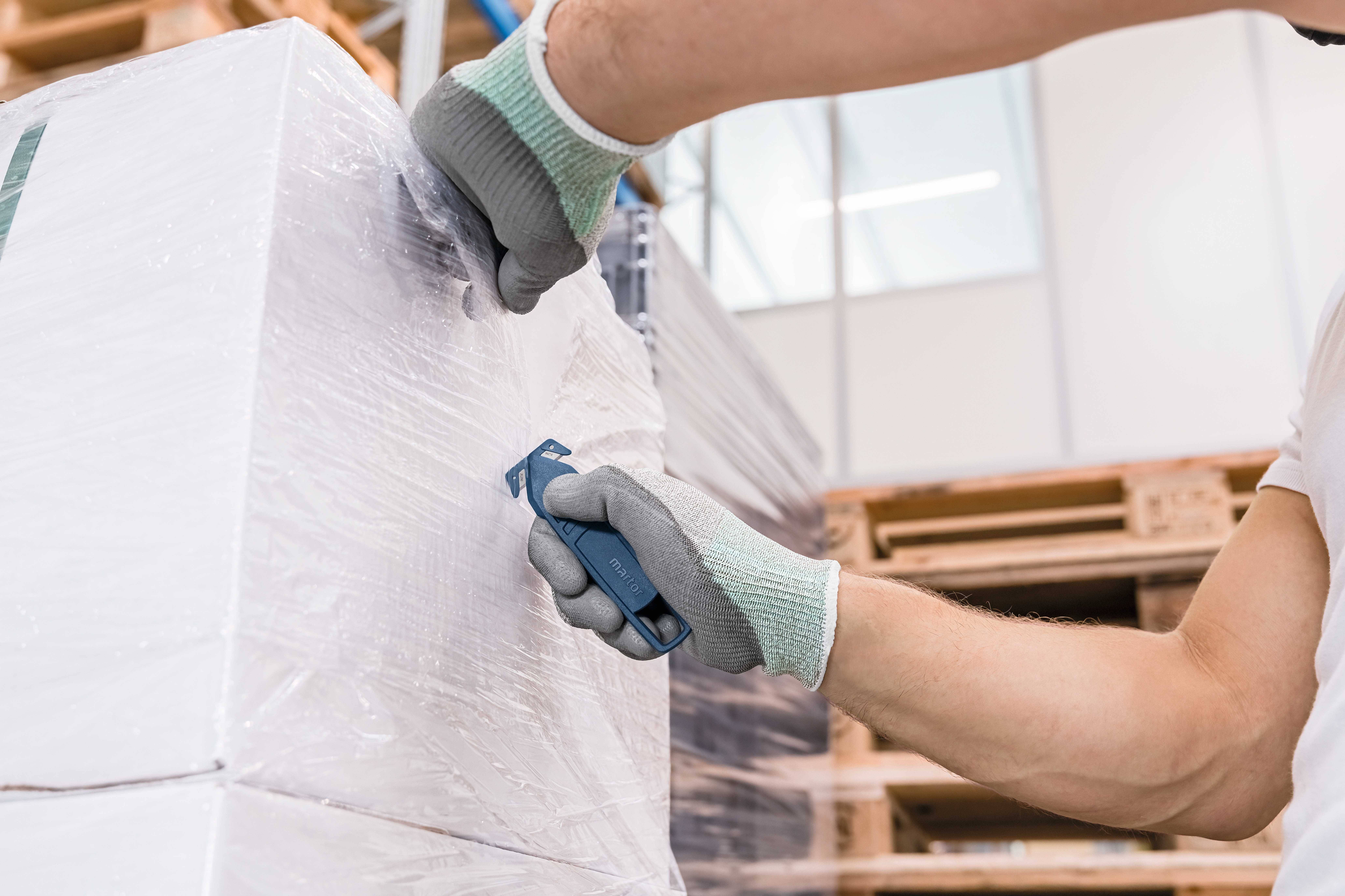 Eine Person mit Handschuhen schneidet Plastikfolie von einer Palette in einem Lager ab. Im Hintergrund sind Regale erkennbar.