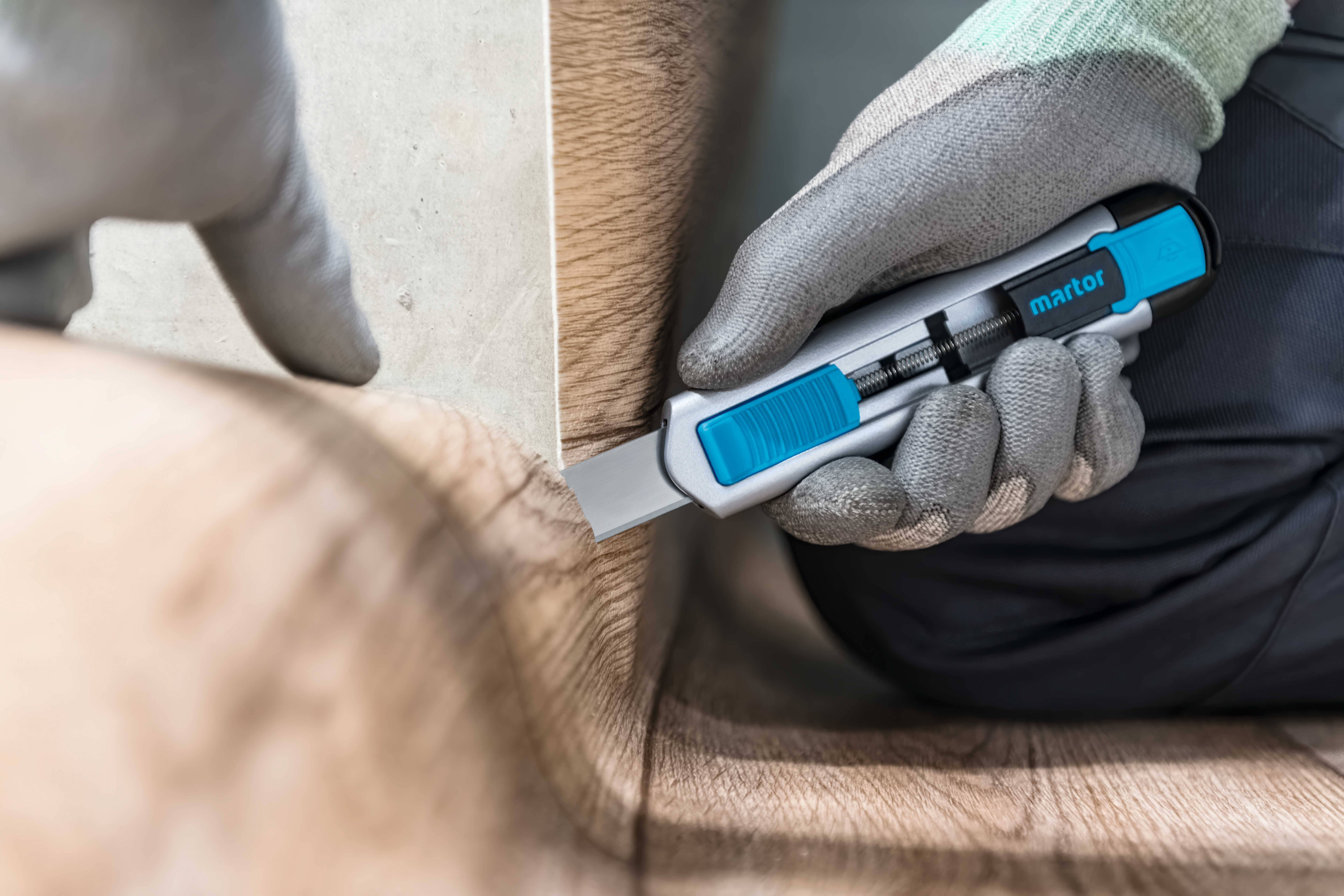 Eine Person trägt Handschuhe und schneidet mit einem blauen Teppichmesser den Rand eines Holzfußbodens sauber entlang einer Kante.