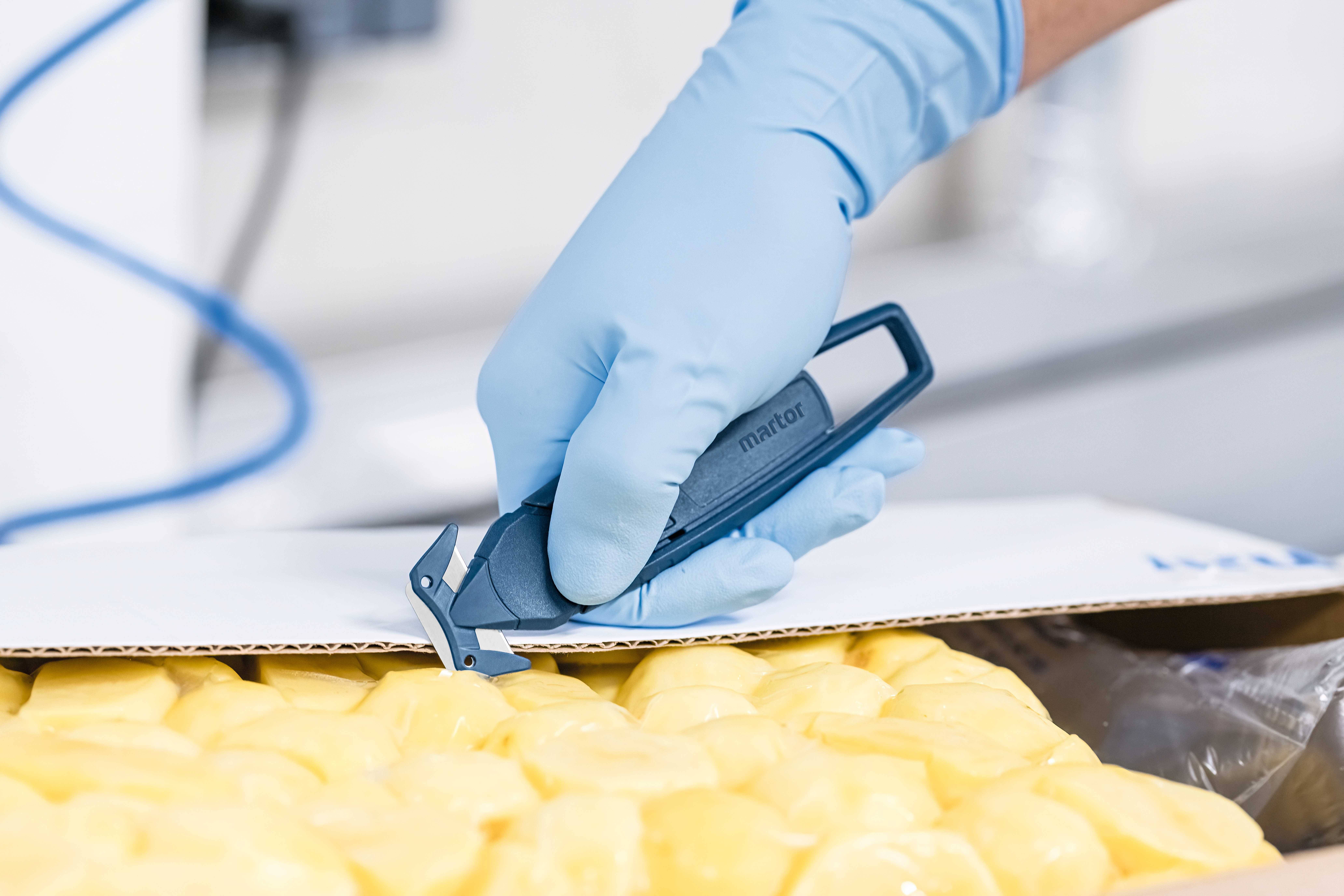 Eine Hand in blauen Handschuhen schneidet mit einem Sicherheitsmesser einen Karton, der mit gelben verpackten Gegenständen gefüllt ist.