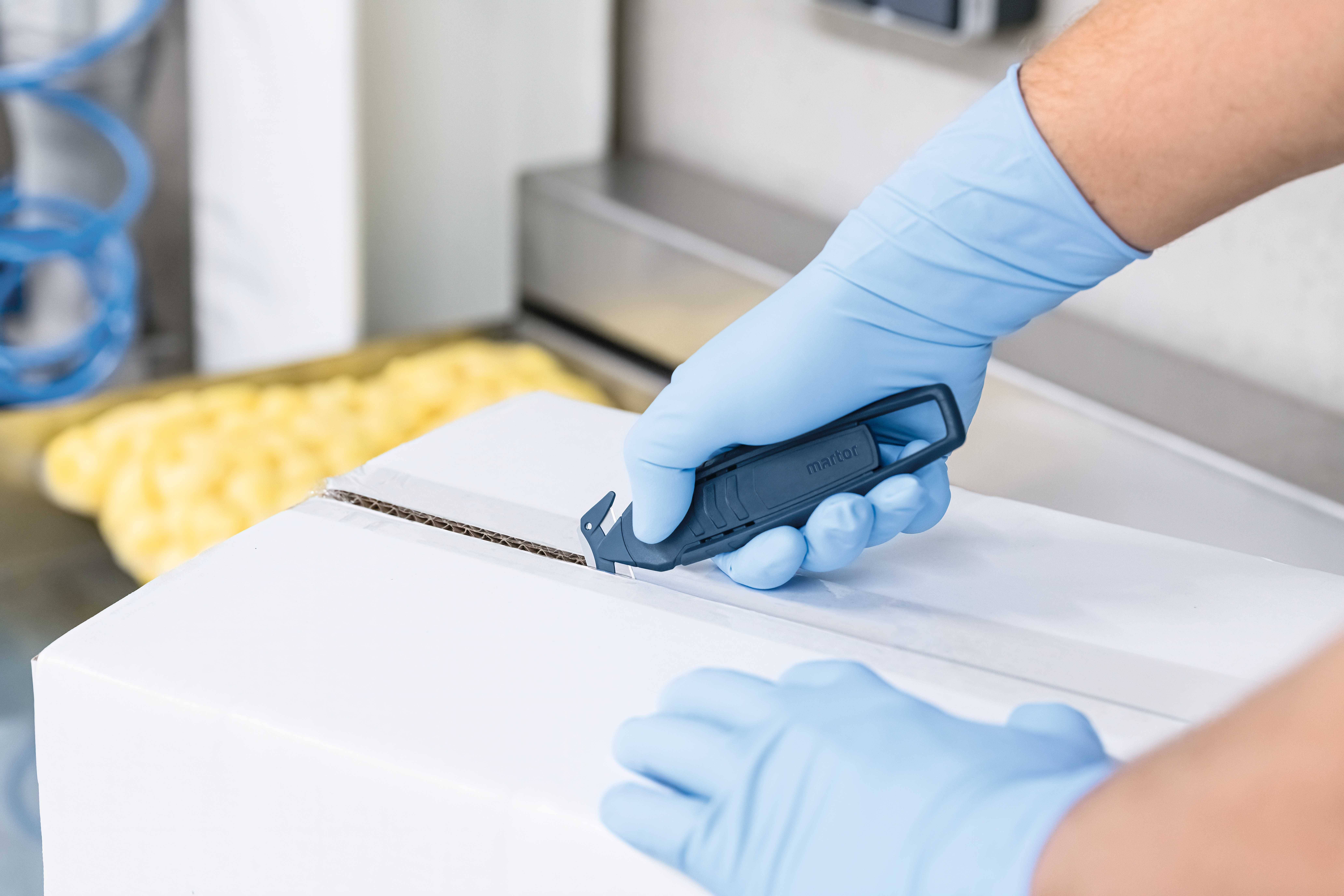 Person mit blauen Handschuhen öffnet mit einem Sicherheitsmesser einen weißen Karton in einer industriellen Umgebung.
