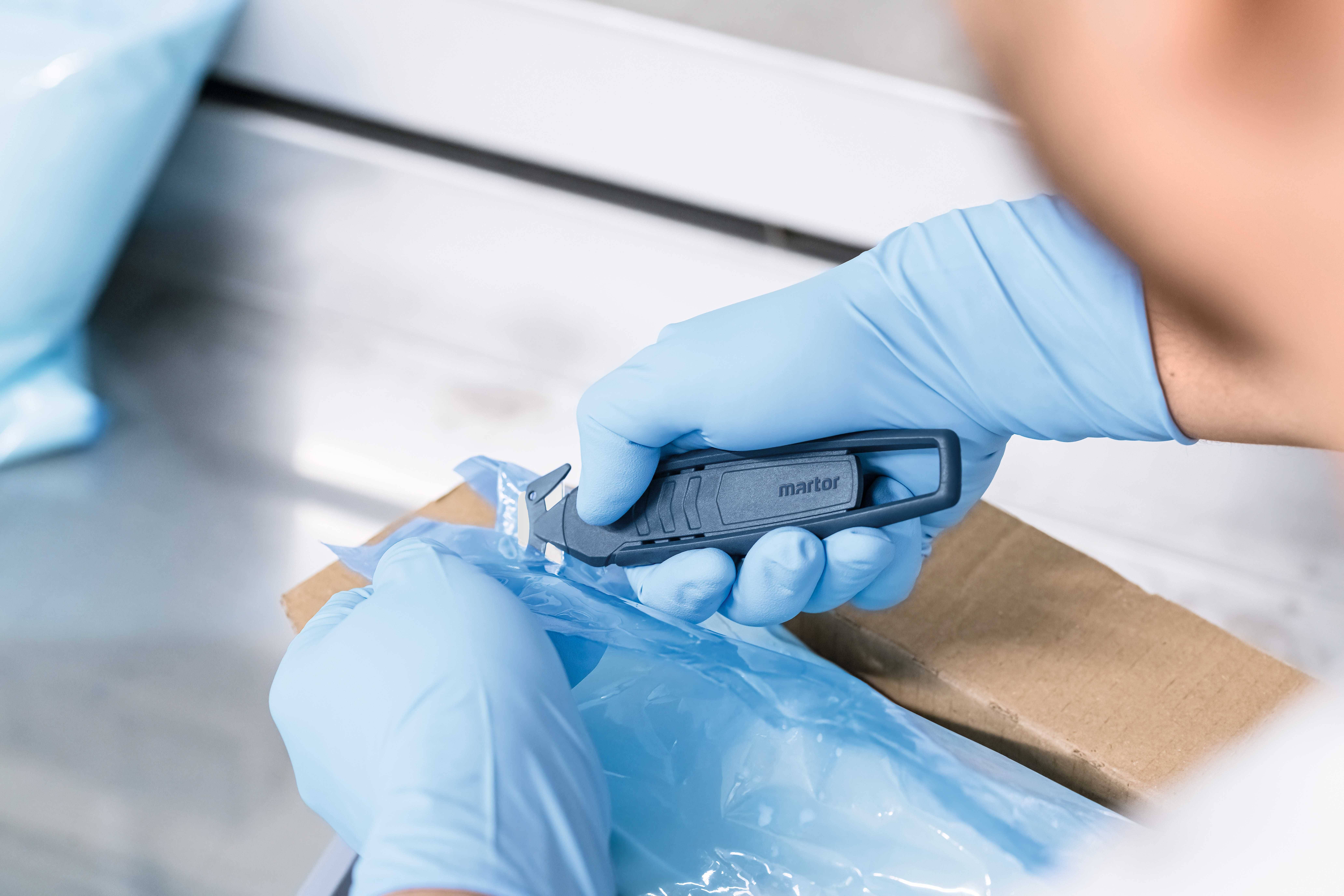 Eine Person mit blauen Handschuhen öffnet vorsichtig eine Verpackung mit einem scharfen Cutter-Messer.