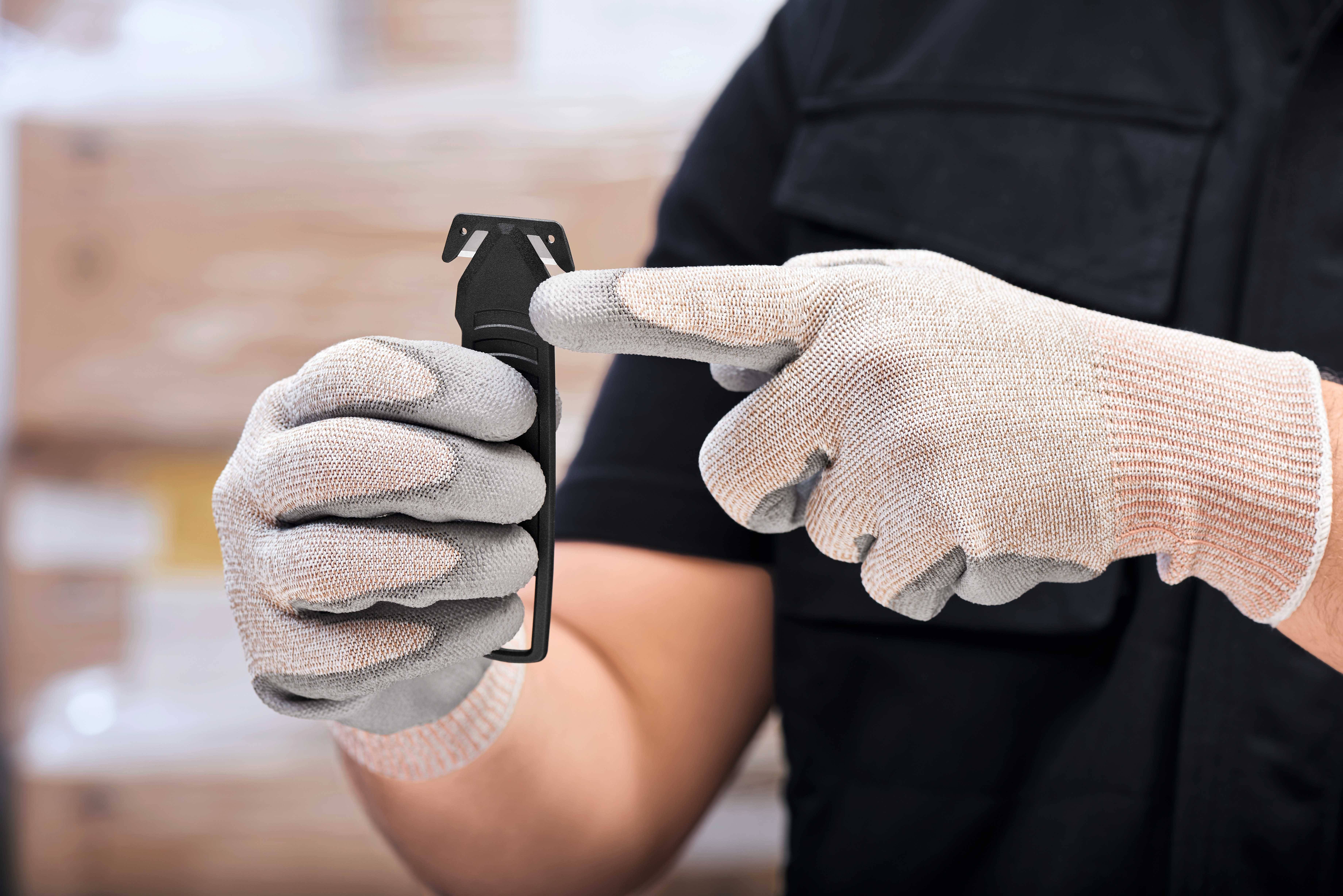 Eine Person trägt graue Handschuhe und zeigt auf einen schwarzen Sicherheitskappenschneider. Hintergrund unscharf, wahrscheinlich Lagerhaus.