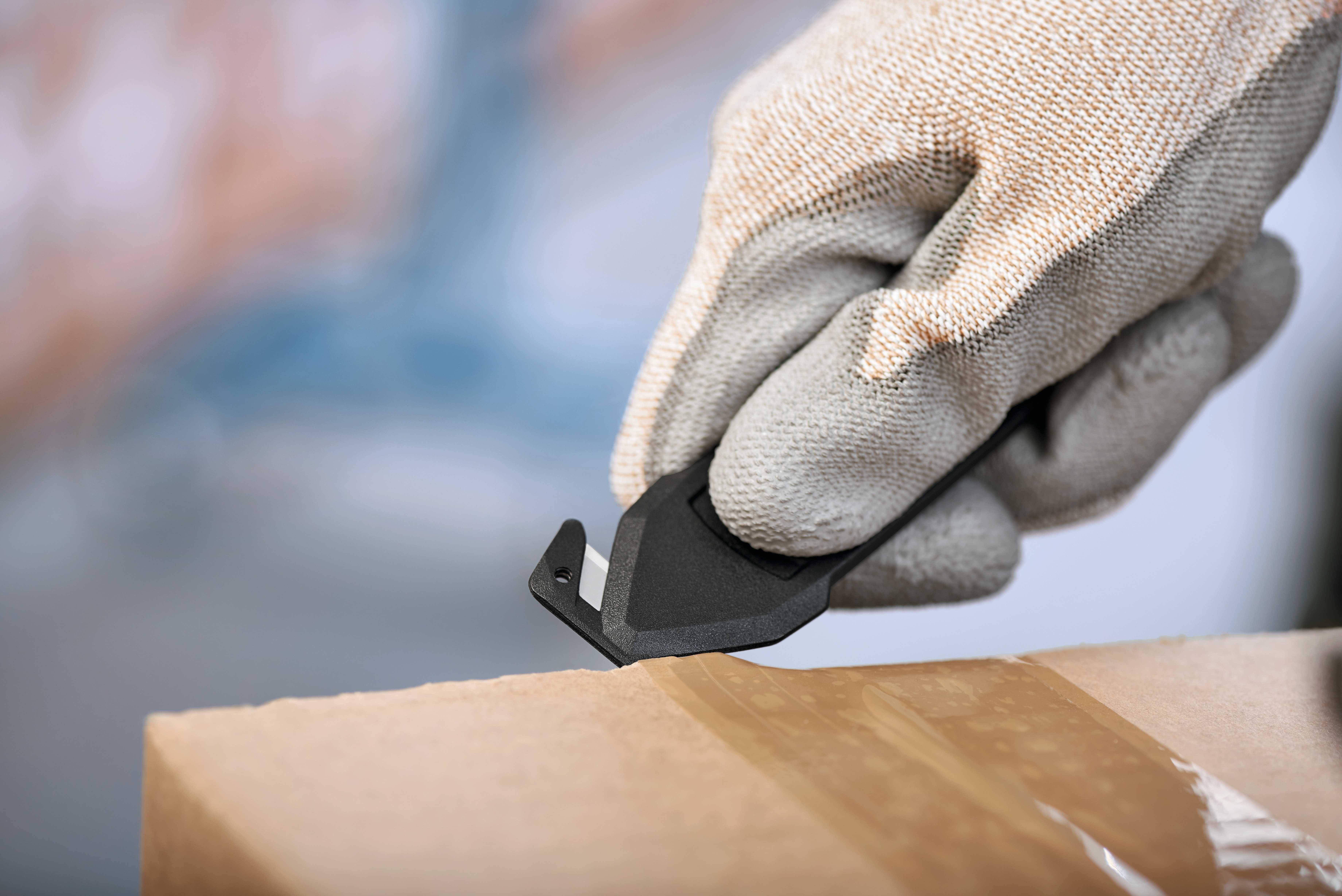 Eine behandschuhte Hand schneidet mit einem Sicherheitsmesser Klebeband auf einem Pappkarton.