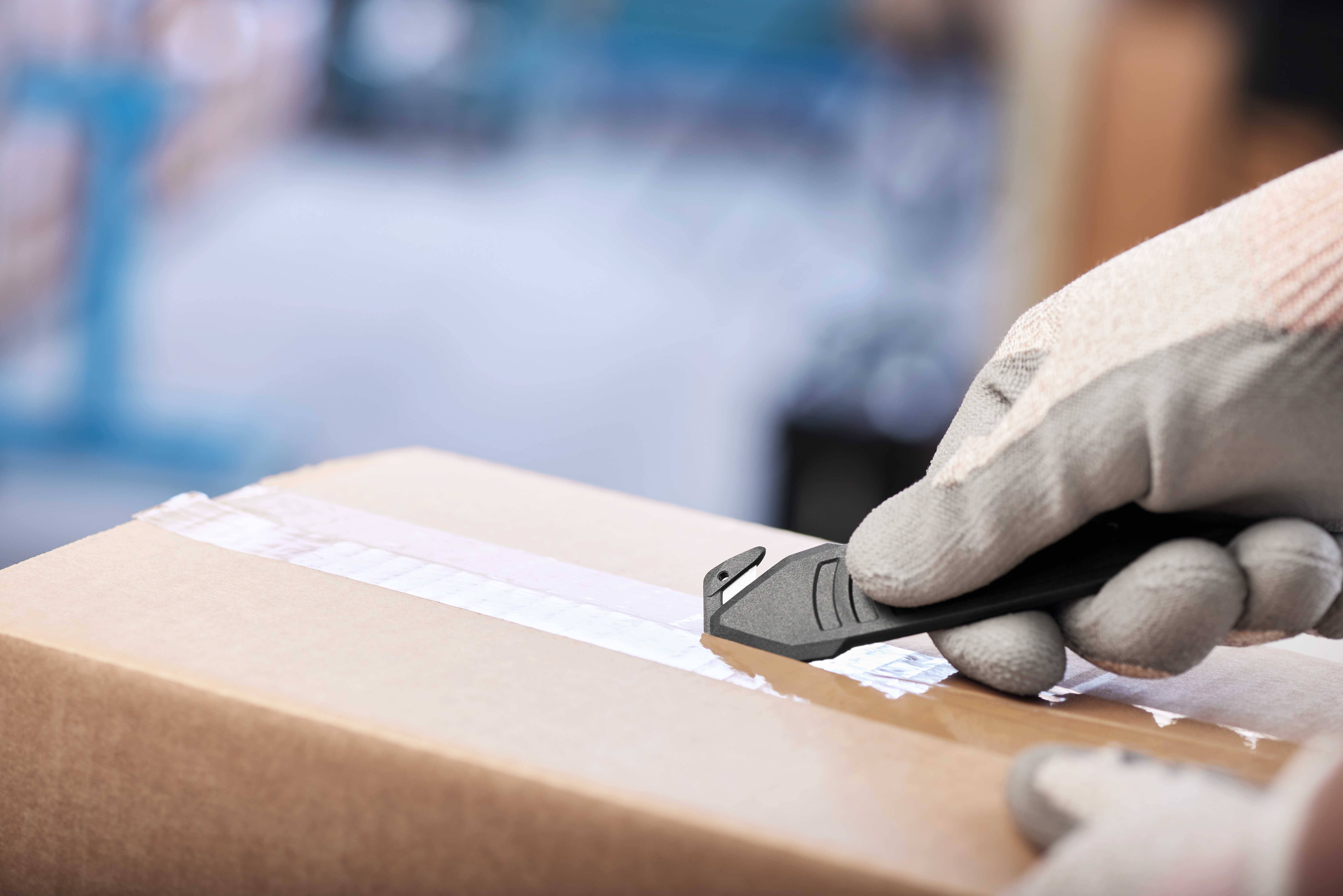 Person schneidet Klebeband auf einem Karton mit Messers. Nahe Fokus auf Hand, deutet auf Paketzustellung oder Verpackung hin.