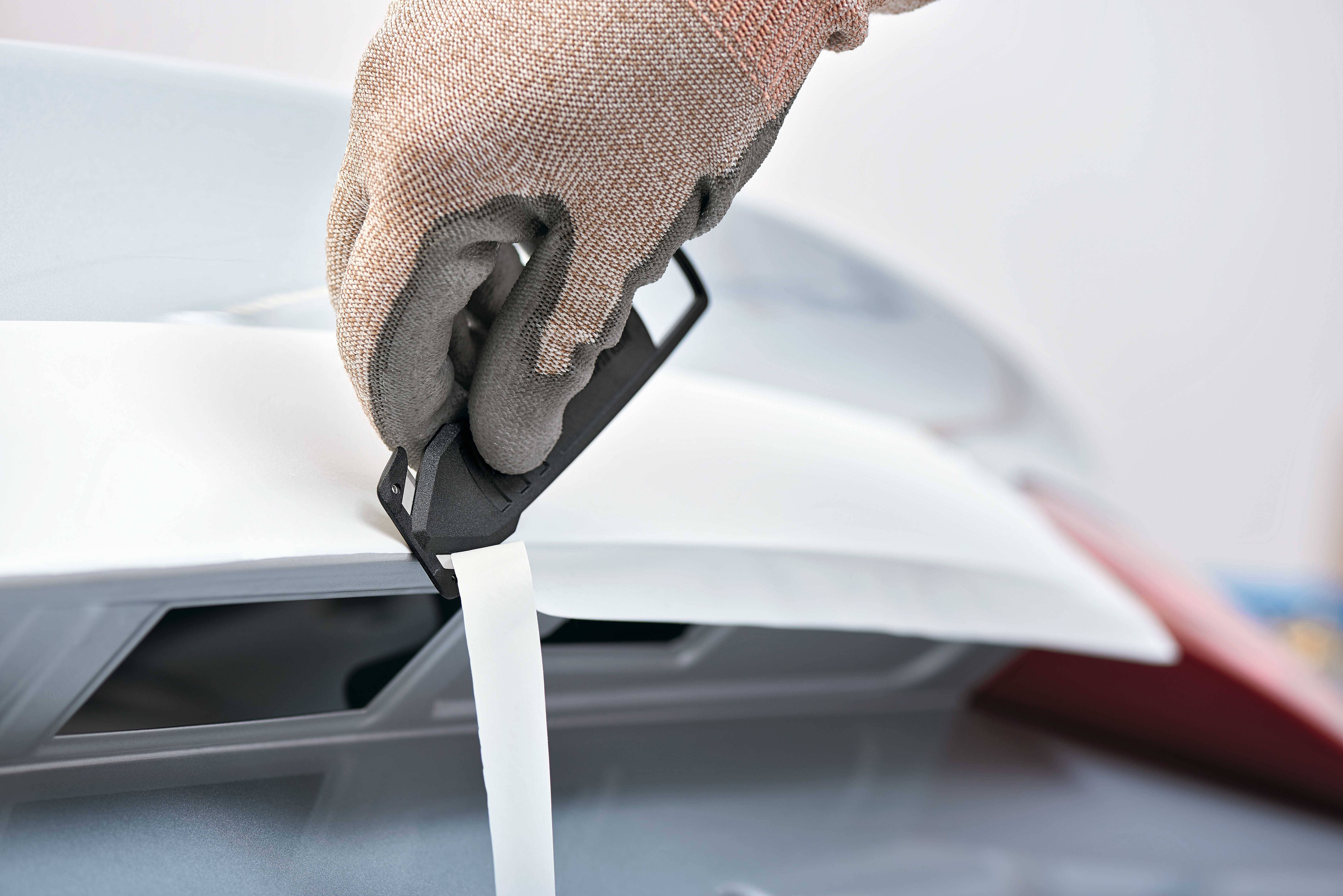 Eine behandschuhte Hand hält einen Kunststoffgriff, der ein elastisches Band spannt, an einem weißen Gehäuse.